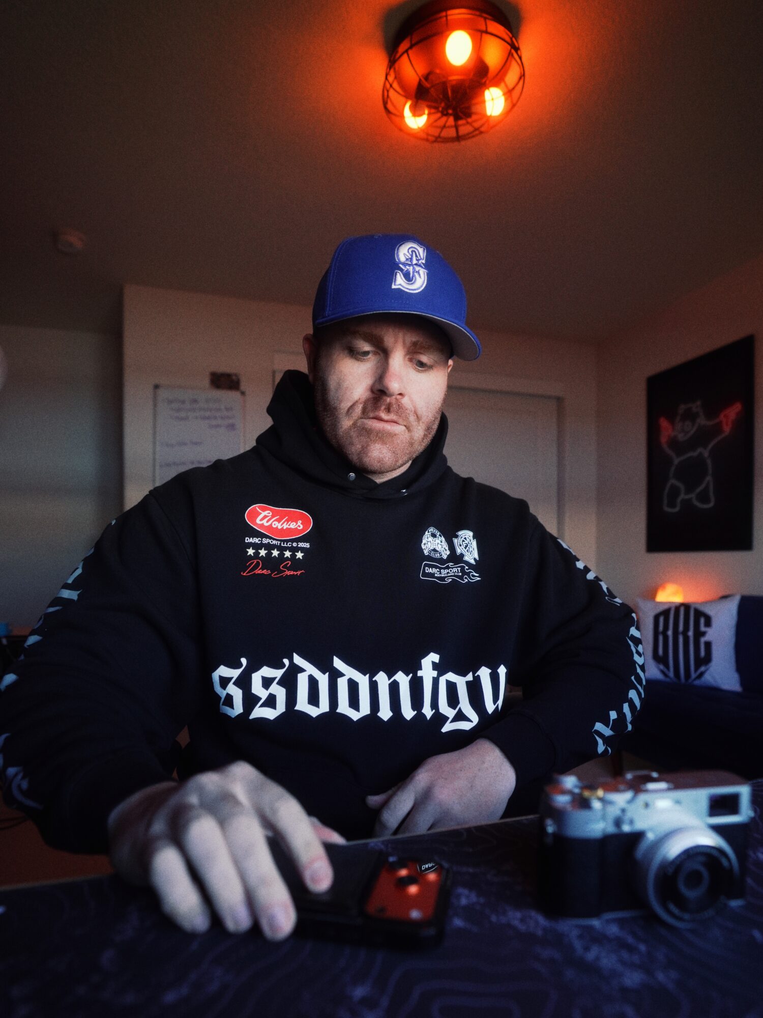 Man wearing a blue cap and black hoodie, sitting at a table with a camera and electronic device, in a dimly lit room.