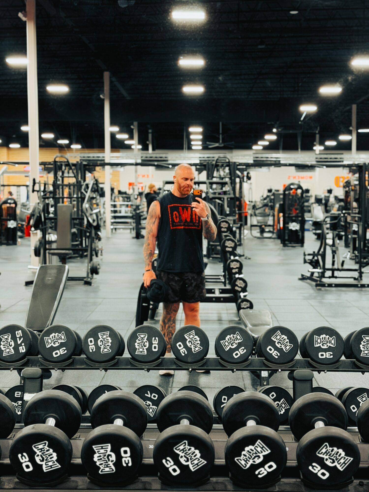 Man taking a selfie in a gym with weights and exercise equipment, bright ceiling lights, and a spacious workout area.