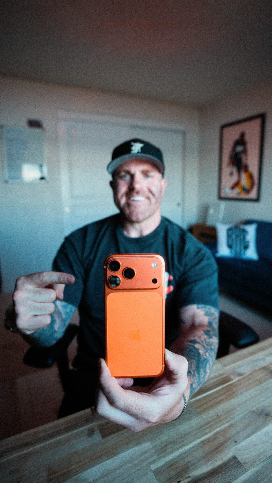 Person taking a selfie with an orange phone, smiling, in a room with a couch and framed artwork on the wall.
