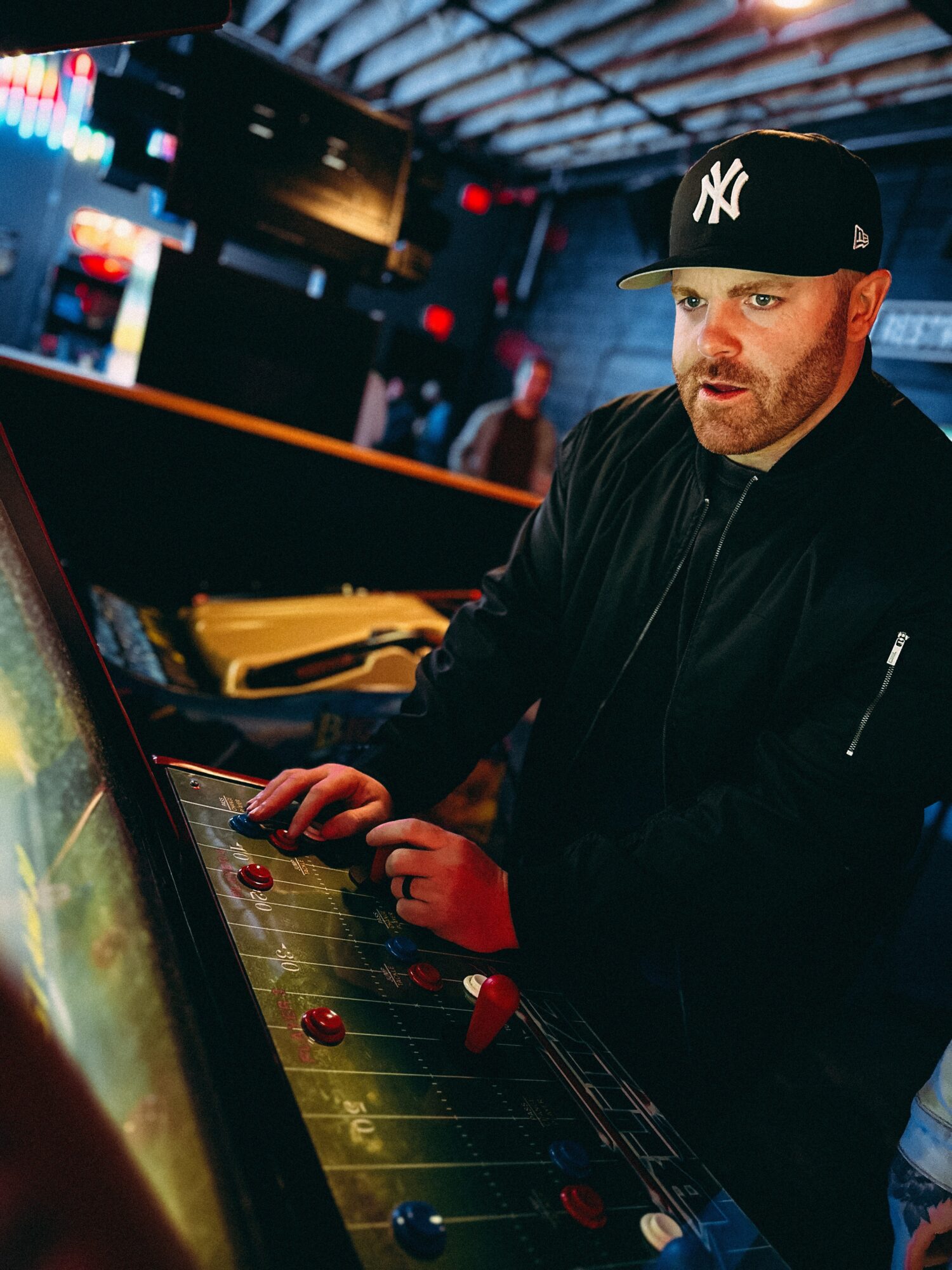 Man playing arcade game, wearing a black jacket and New York Yankees cap, in a dimly lit arcade setting.