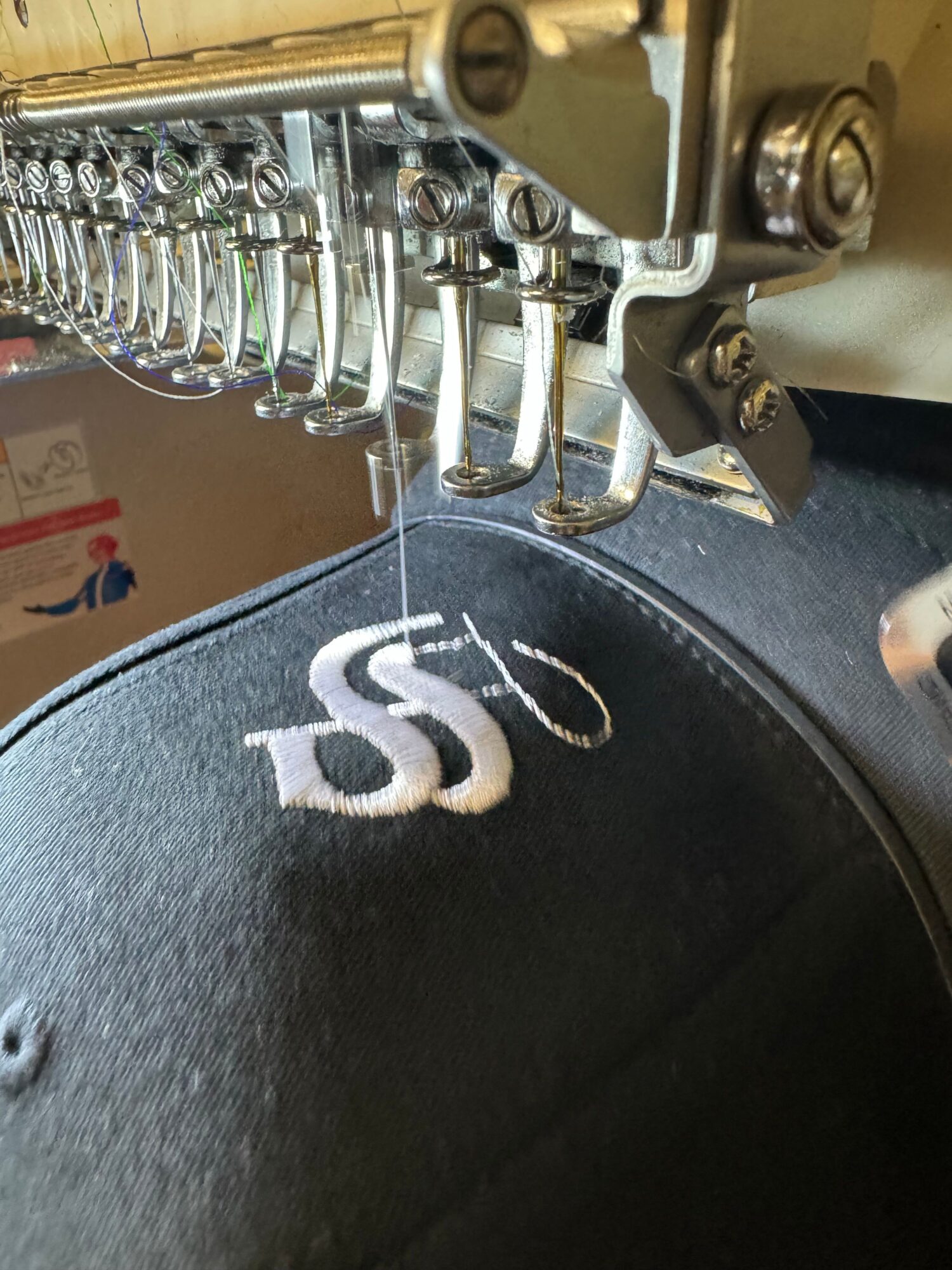 Close-up of embroidery machine stitching white thread on black fabric with a partially visible logo.