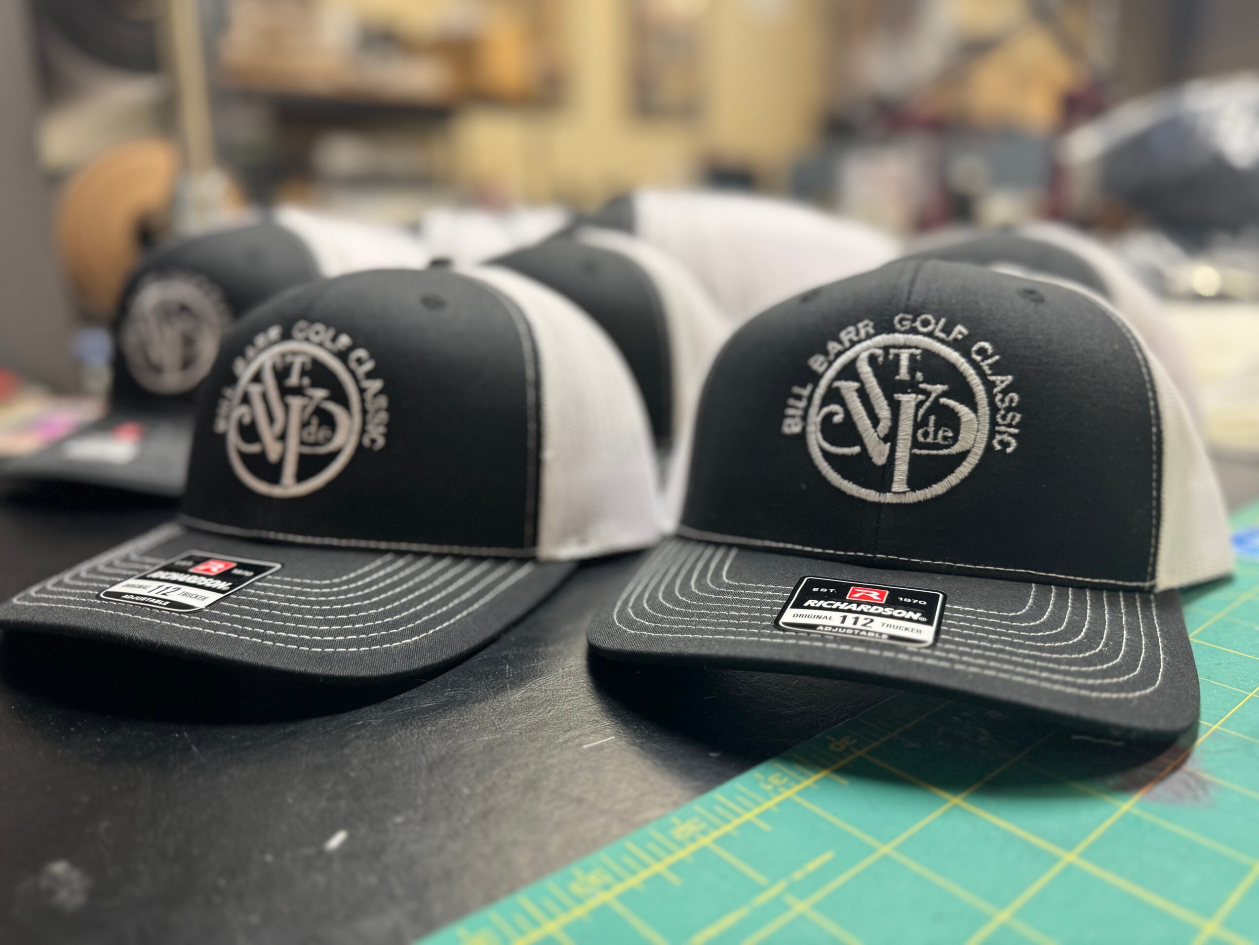 Six black and white trucker hats with logo on front, placed on a table with a cutting mat, in a workshop setting.
