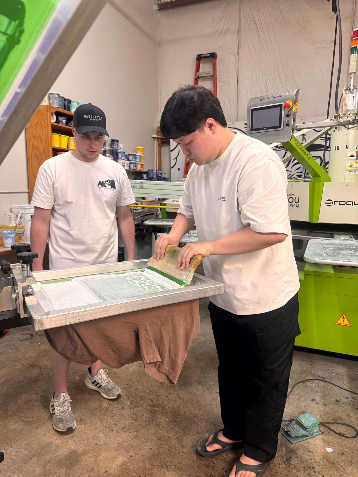 Two people working at a table in a workshop or factory setting, with machinery and equipment around them.