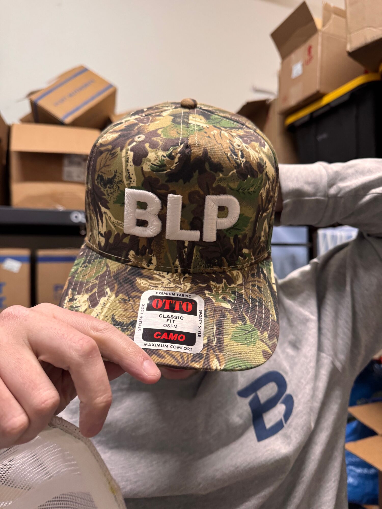 Person holding camouflage BLP cap with patch, in storage room with boxes and shelves in background.