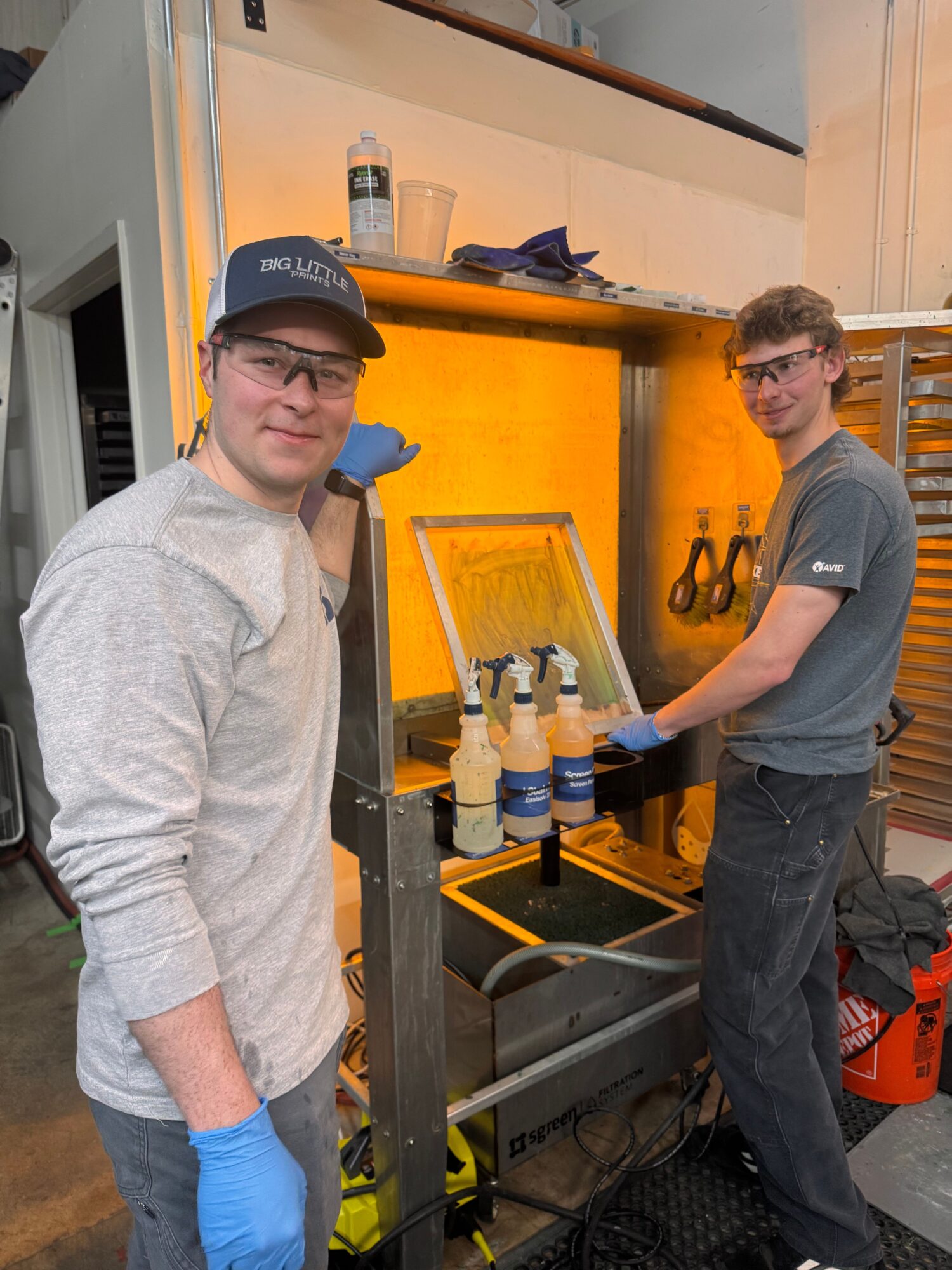Two people working with a yellow machine, one wearing a cap and gloves, in a workshop setting.