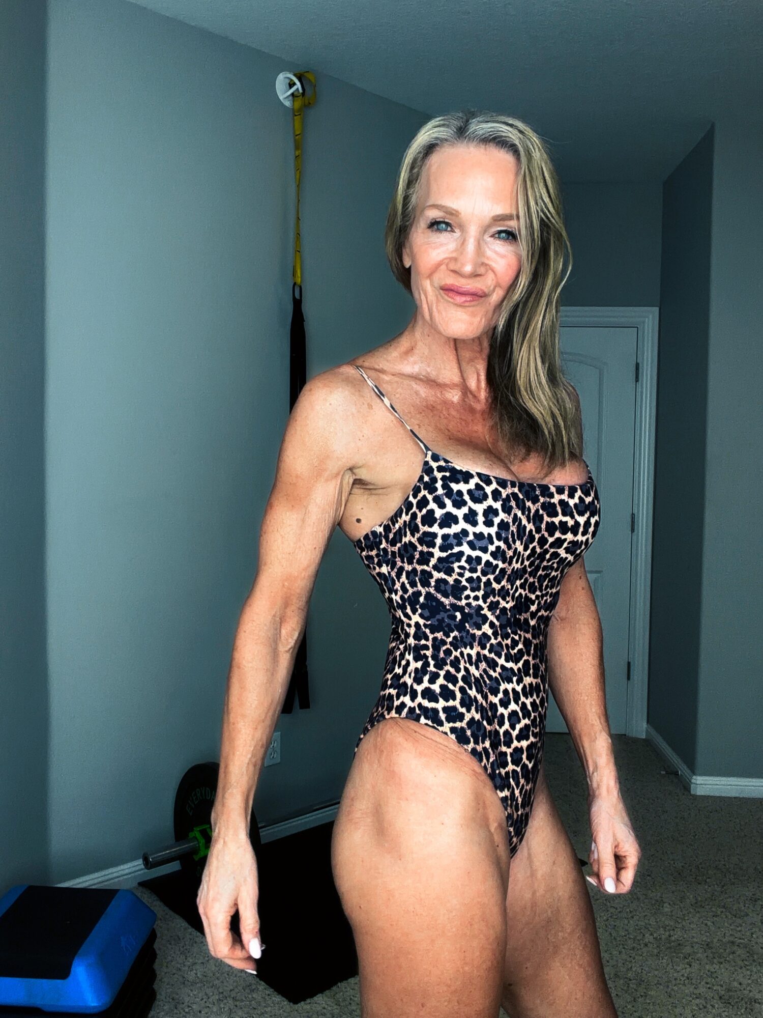 A woman with long hair in a leopard print swimsuit standing in a room with gray walls and a door in the background.