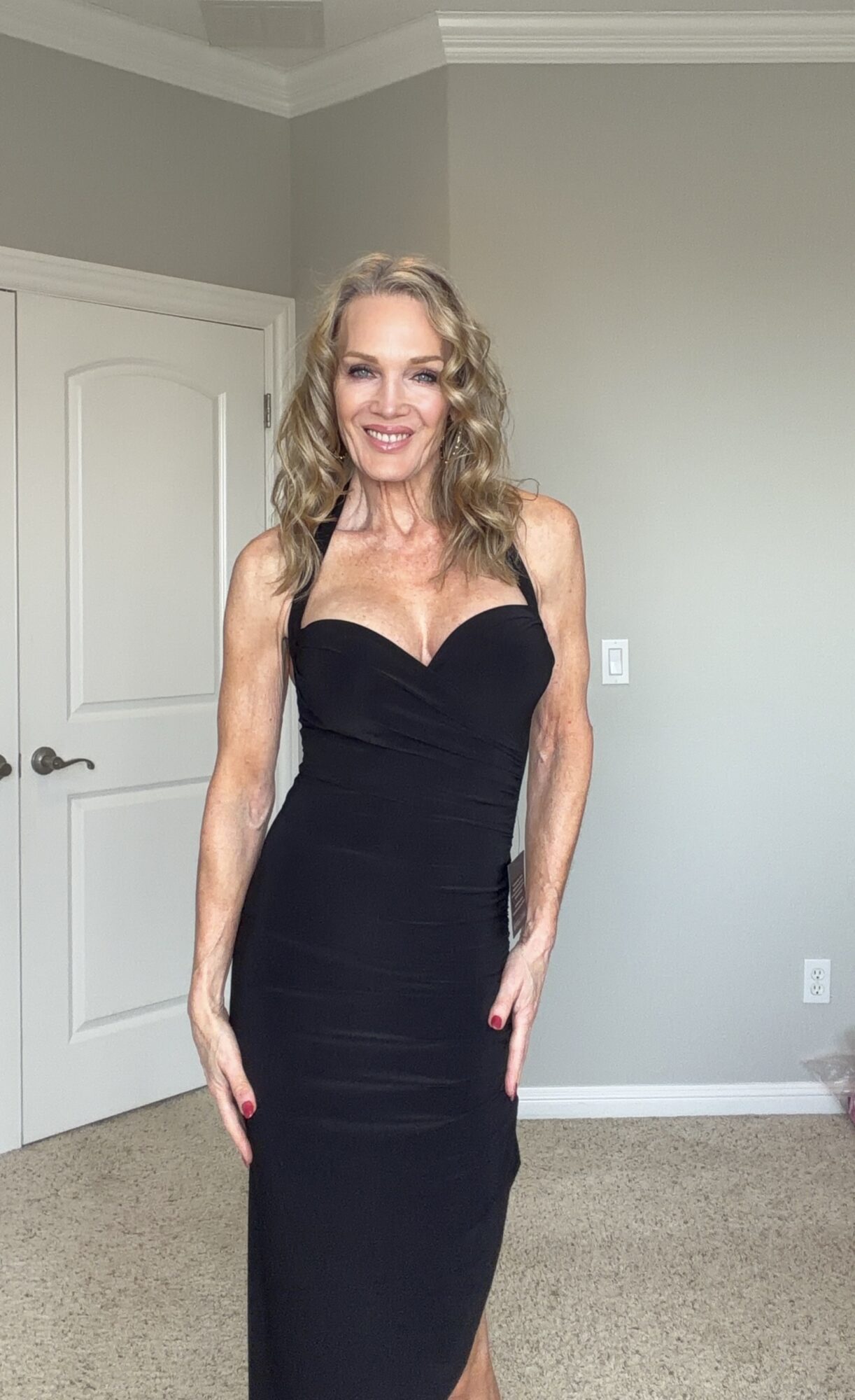 Woman with wavy blonde hair in a black strapless dress smiling in a room with gray walls and a white door.