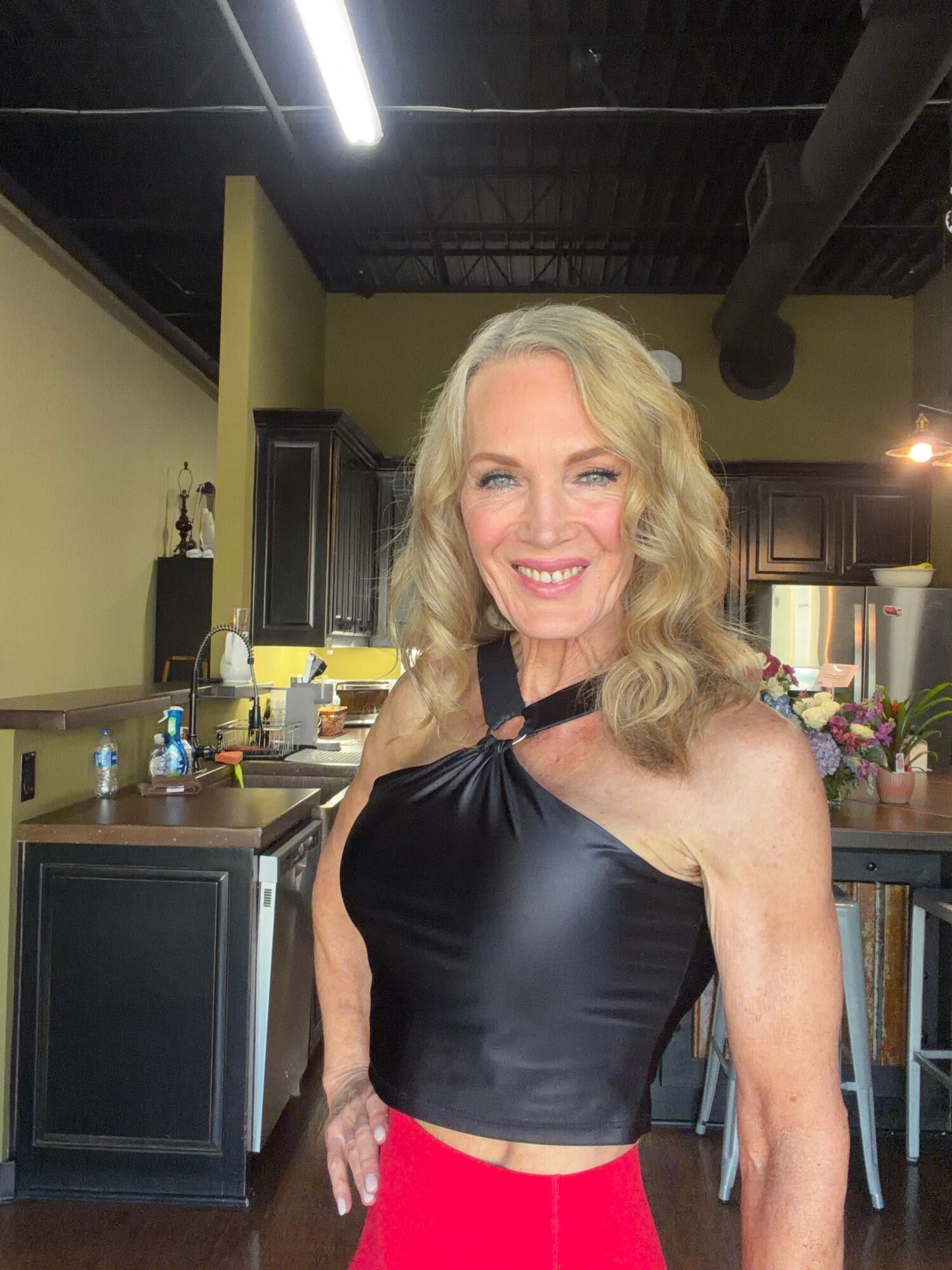 Smiling woman with wavy blonde hair in a black sleeveless top and red skirt in a kitchen setting.