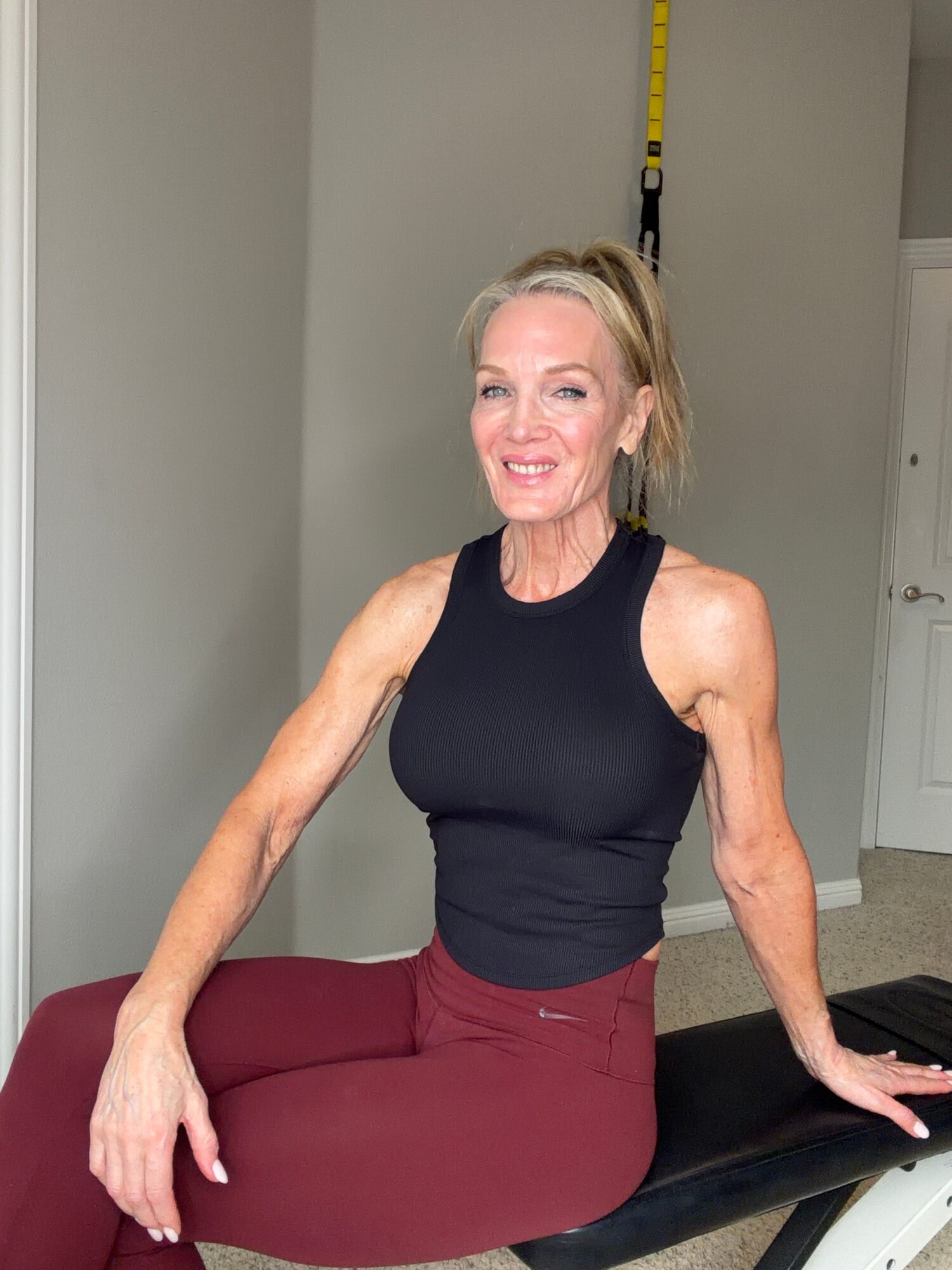 Woman in workout attire sitting on a bench in a fitness room, smiling, with a yellow strap hanging on the wall behind her.
