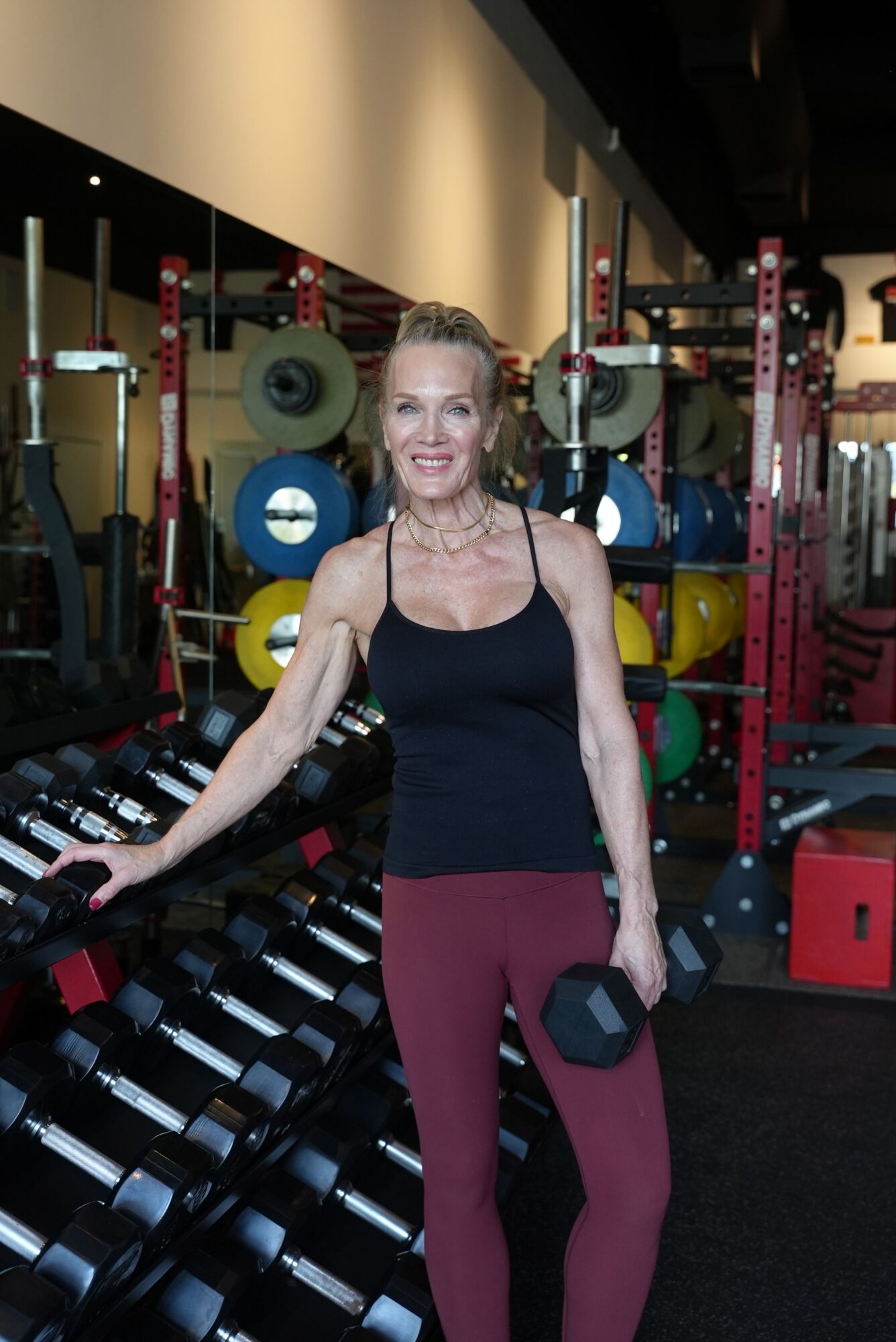 Woman in workout clothes holding a dumbbell in a gym with weights and equipment in background.