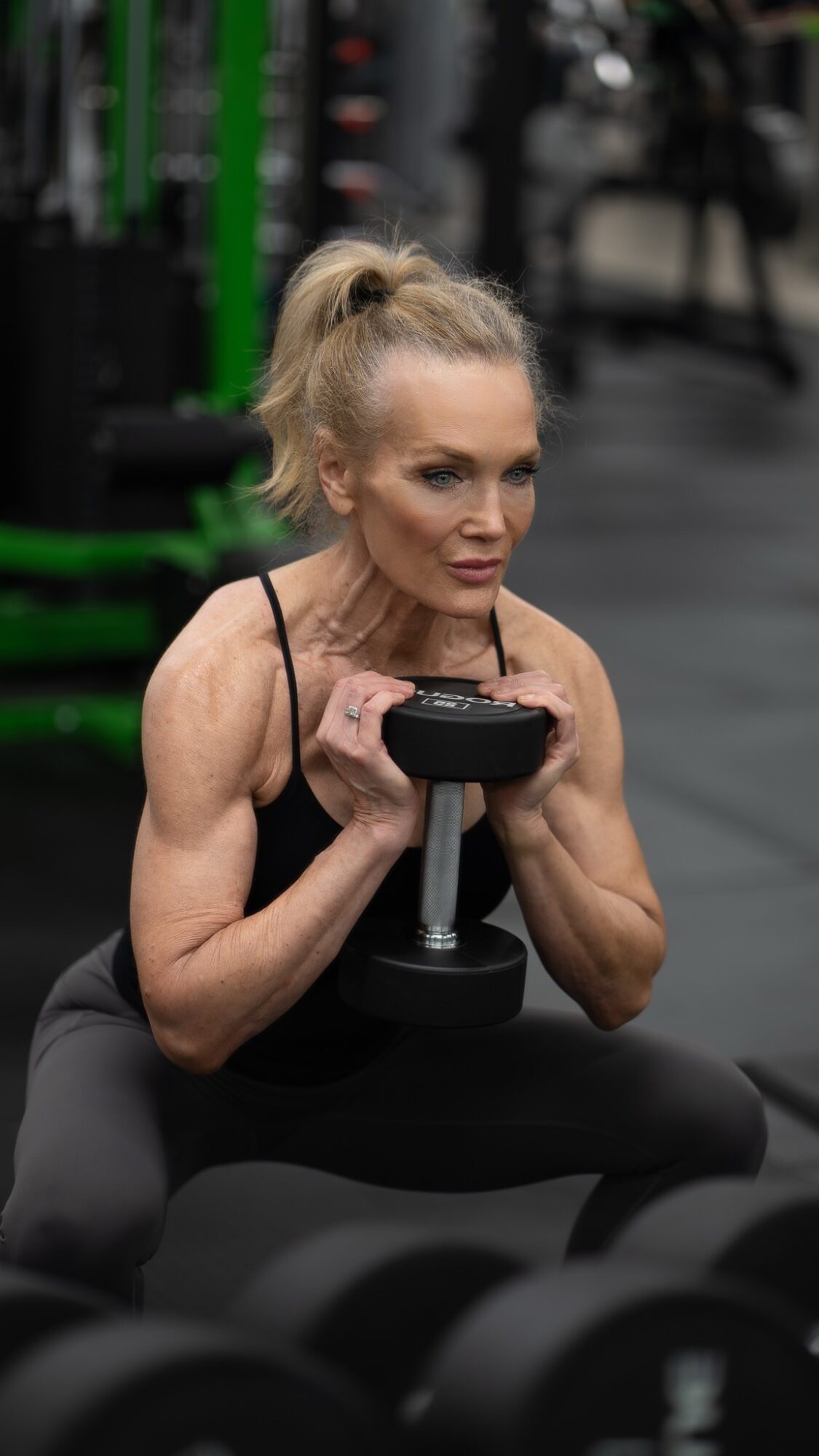 Woman with blonde hair in a ponytail lifting a dumbbell in a gym, wearing a black tank top.