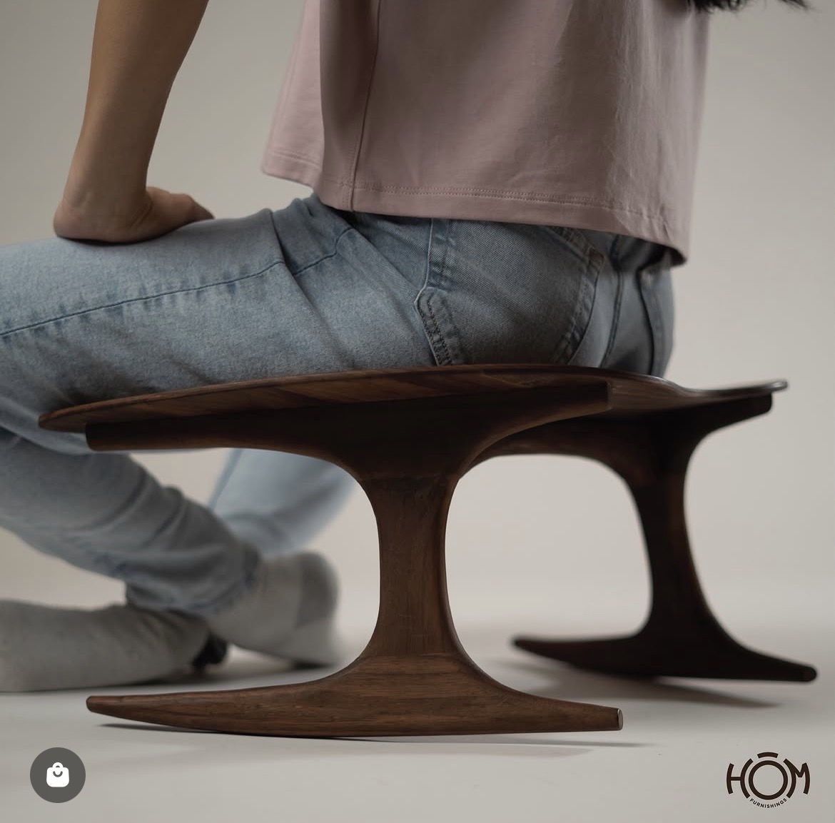 Person sitting on a wooden bench with a curved design, wearing jeans and a pink top, against a plain background.