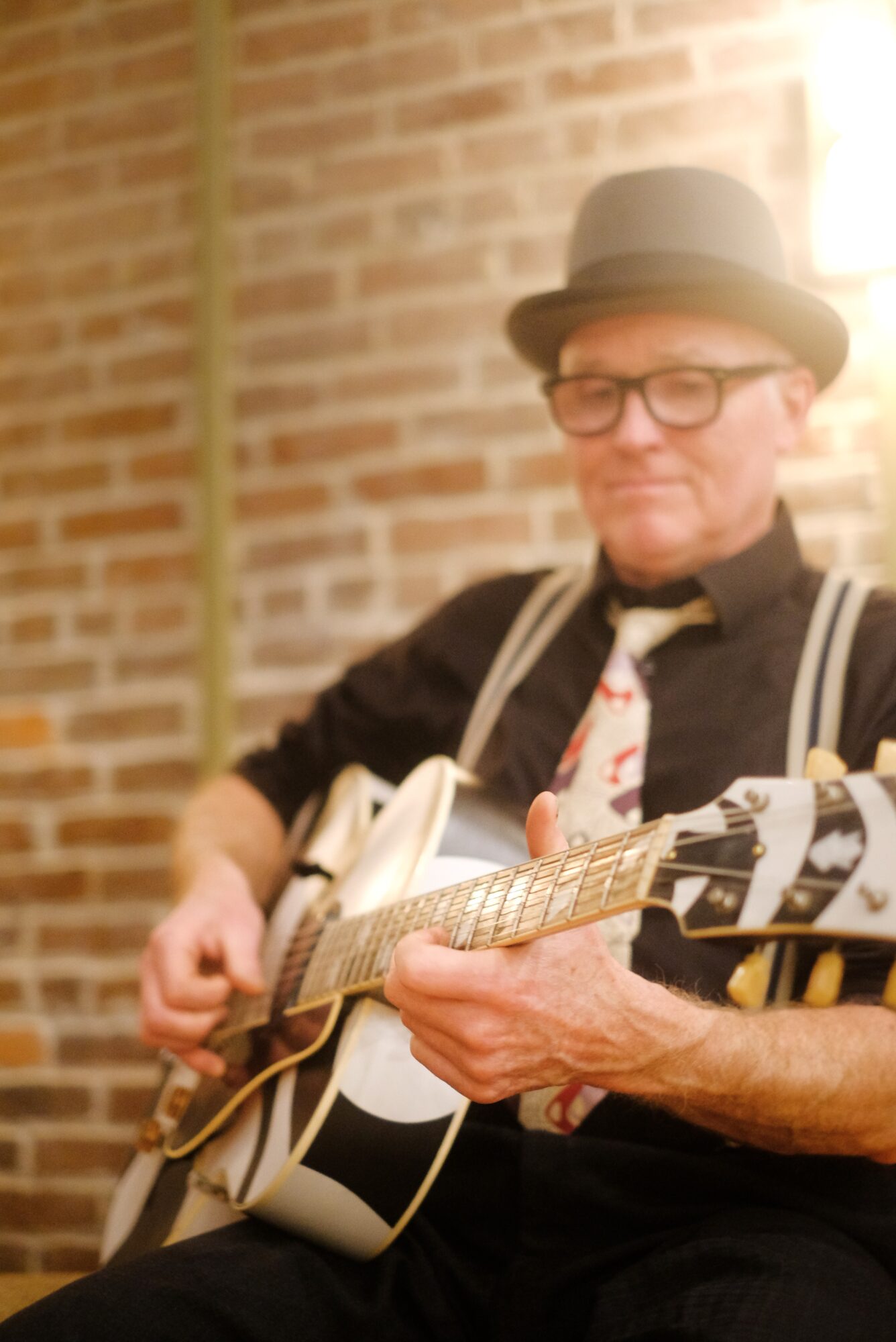Person wearing glasses and a hat playing an acoustic guitar against a brick wall.