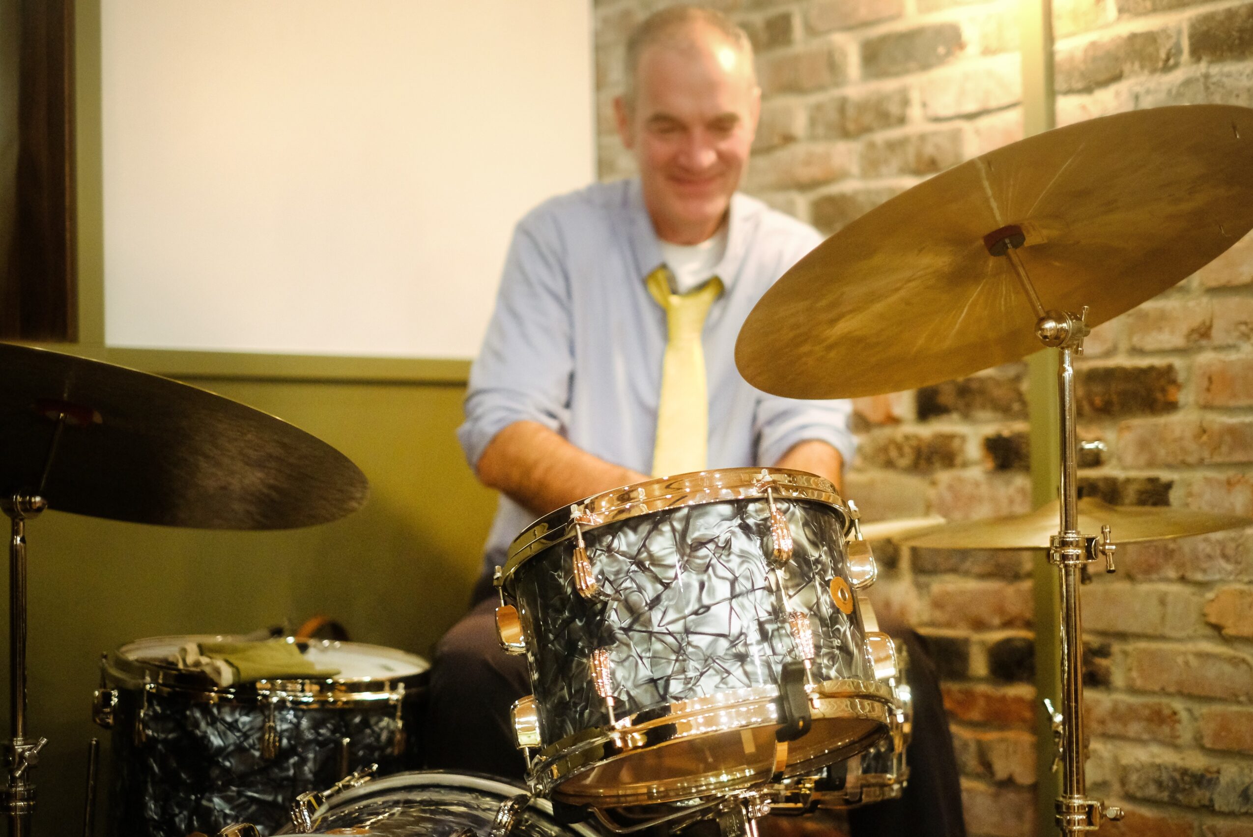 Man smiling behind a drum set in a room with brick wall, wearing a light shirt and yellow tie.