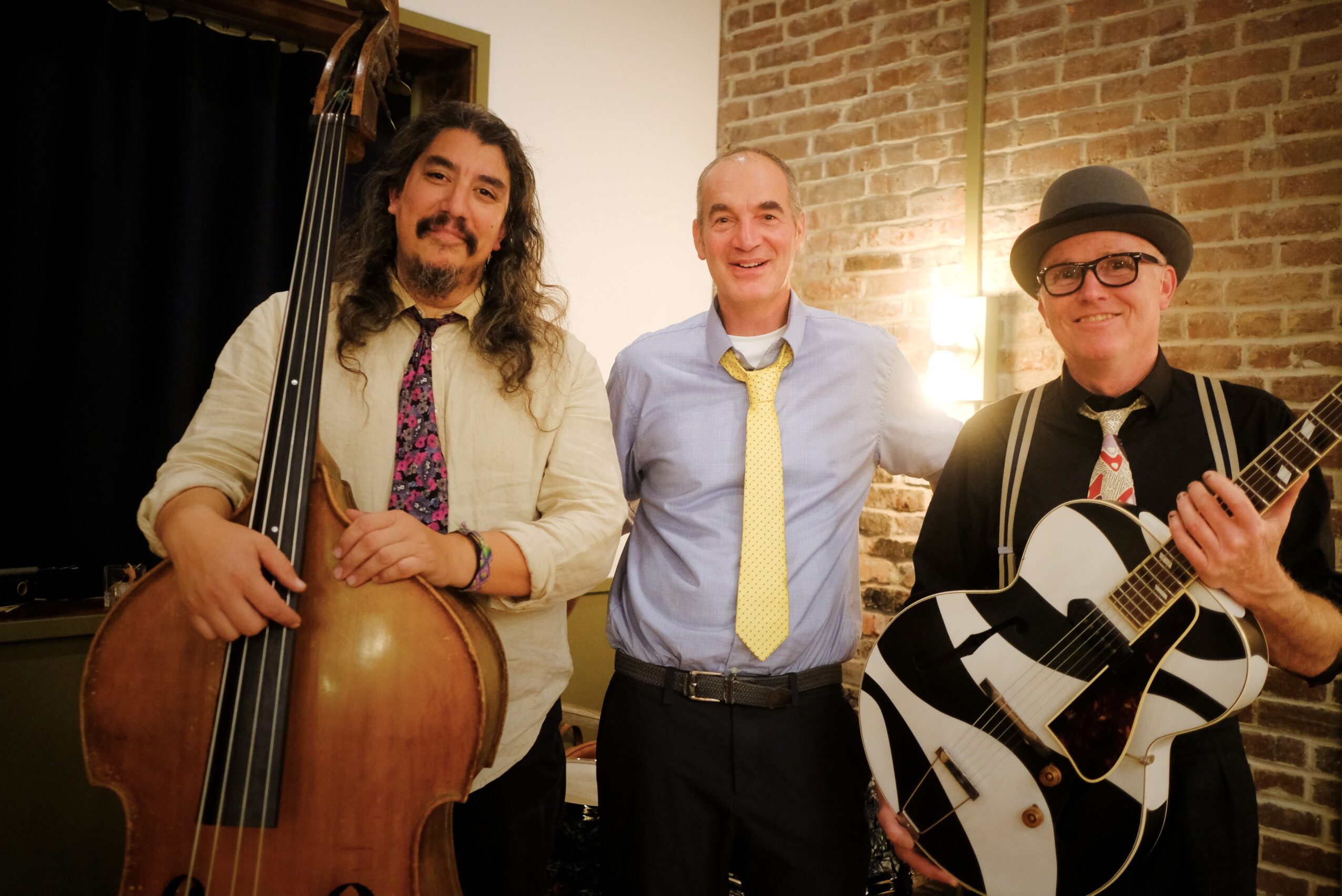 Three men standing indoors, one holding a double bass, another with a guitar, and the third smiling in front of a brick wall.