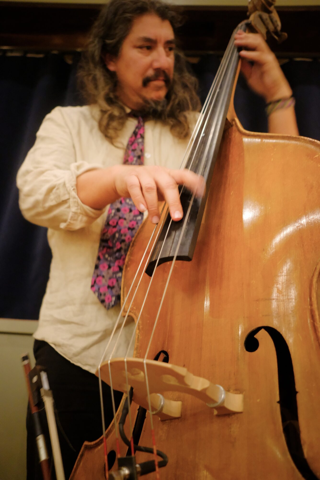 Person playing double bass, focusing on their hands and instrument, with long hair and a mustache, wearing a light shirt and colorful tie.