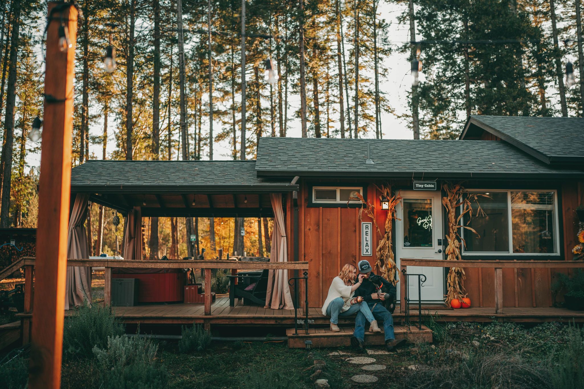 Wooden cabin with porch and trees in background, two people sitting on steps, autumn setting