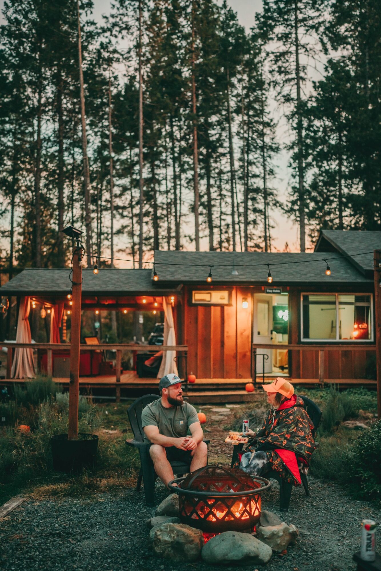 Two people sit around a campfire outside a wooden cabin with string lights, trees in the background, evening setting.