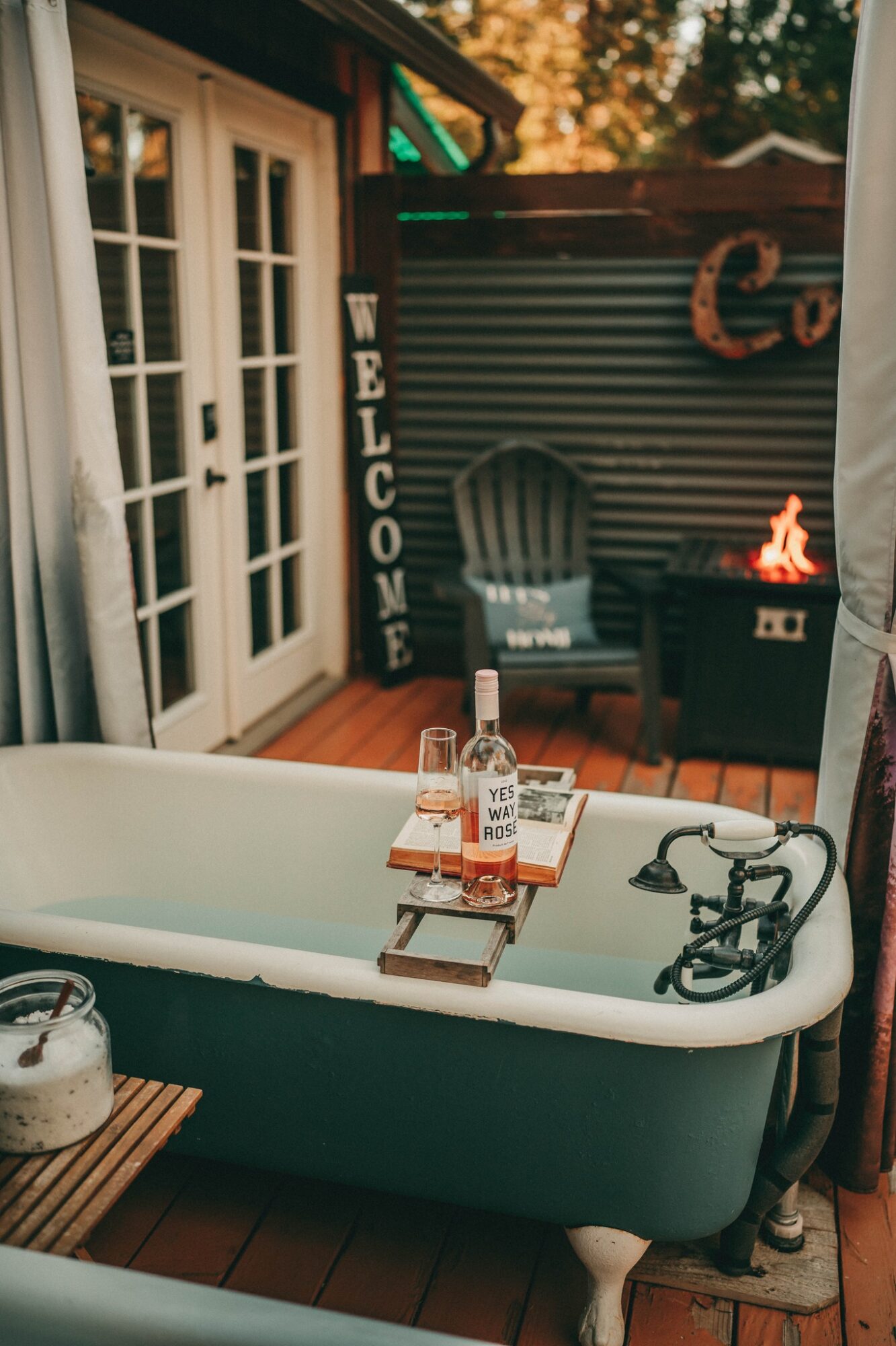 Outdoor patio with a bathtub, a welcome sign, a chair, and a small fire pit, surrounded by trees and a building with glass doors.