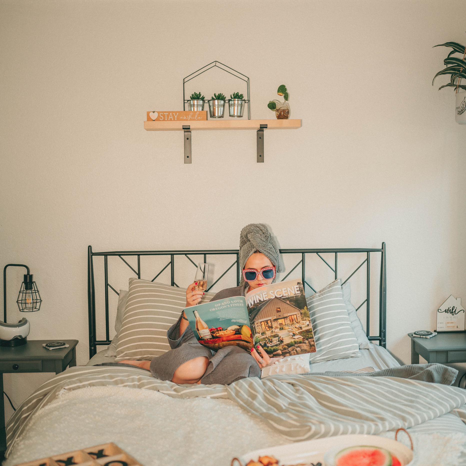 Child with towel on head and sunglasses reading a book in bed, with pillows and nightstands on either side, and wall decor above.