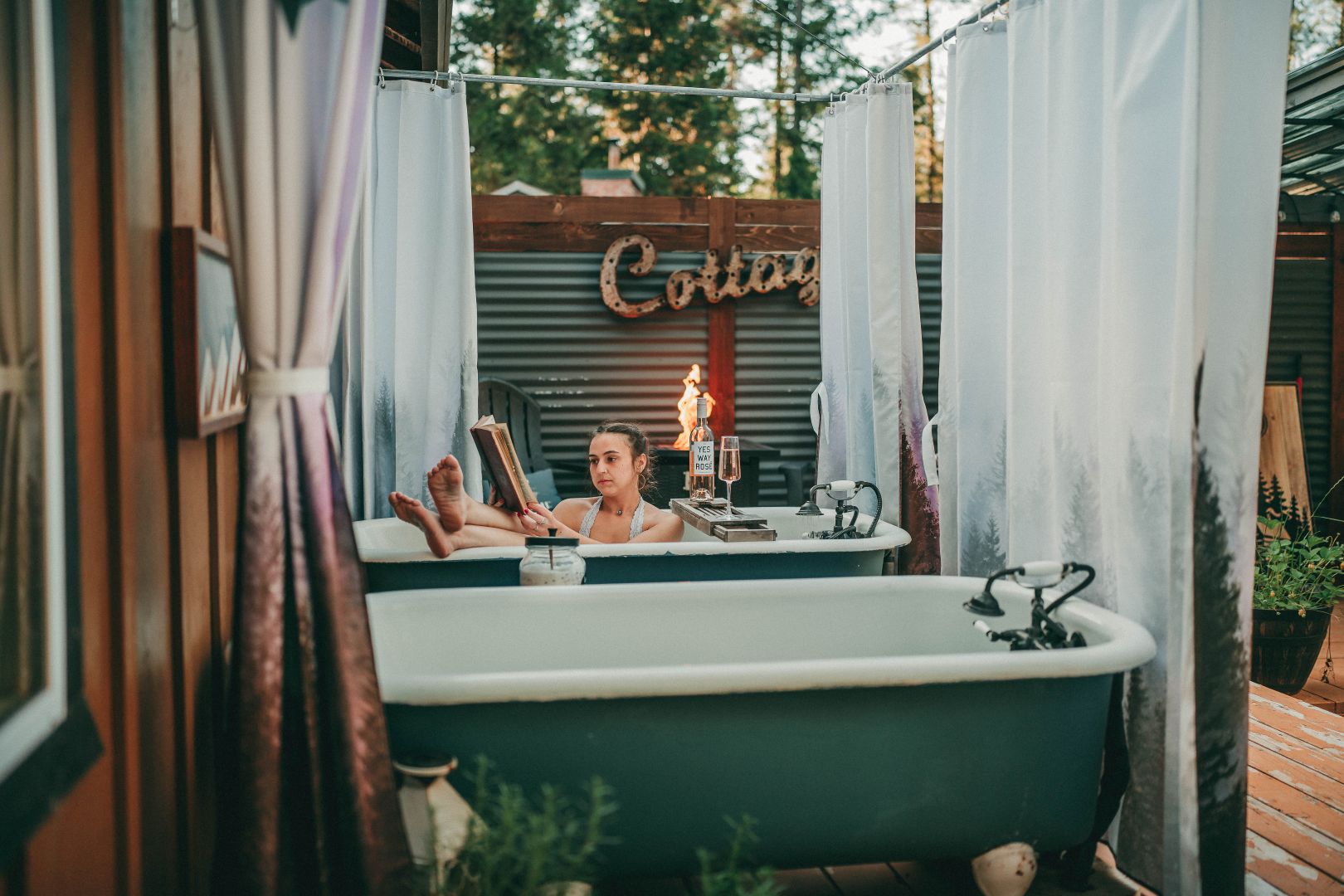 Person relaxing in a bathtub outdoors with curtains, trees, and a sign in the background.