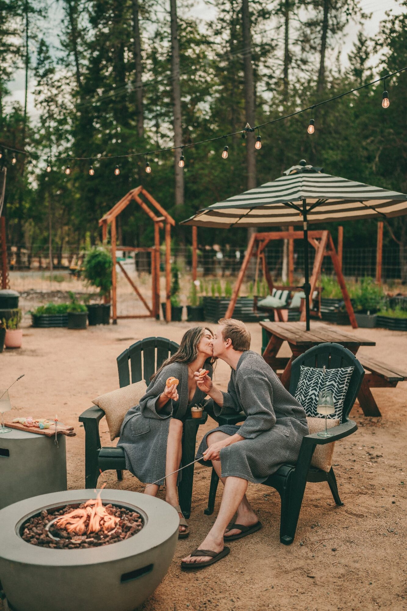 Two people sit on outdoor chairs near a fire pit, talking and smiling, with a playground and string lights in the background.