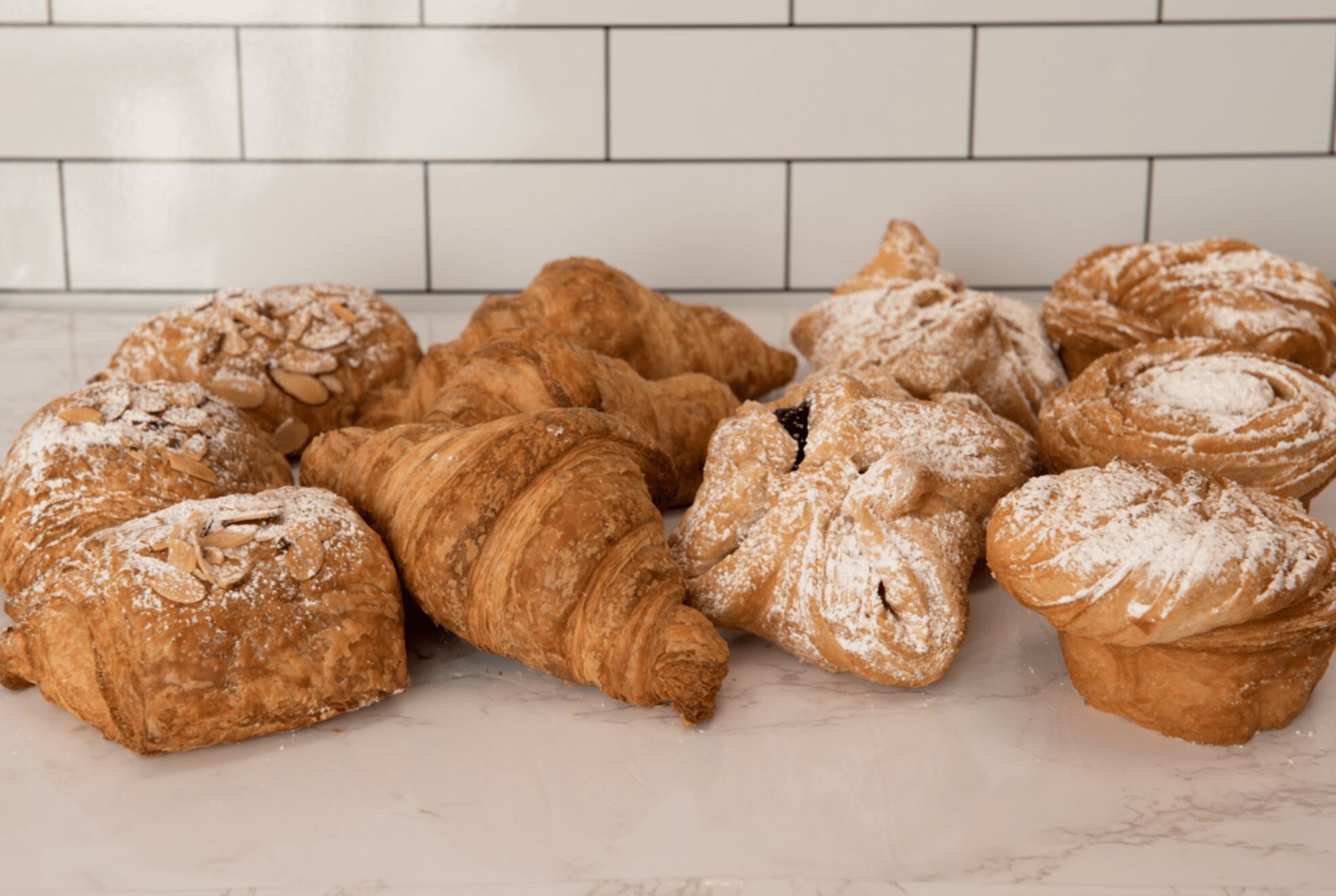 Assorted pastries including croissants, muffins, and dusted pastries on a marble surface with a white tiled wall background.
