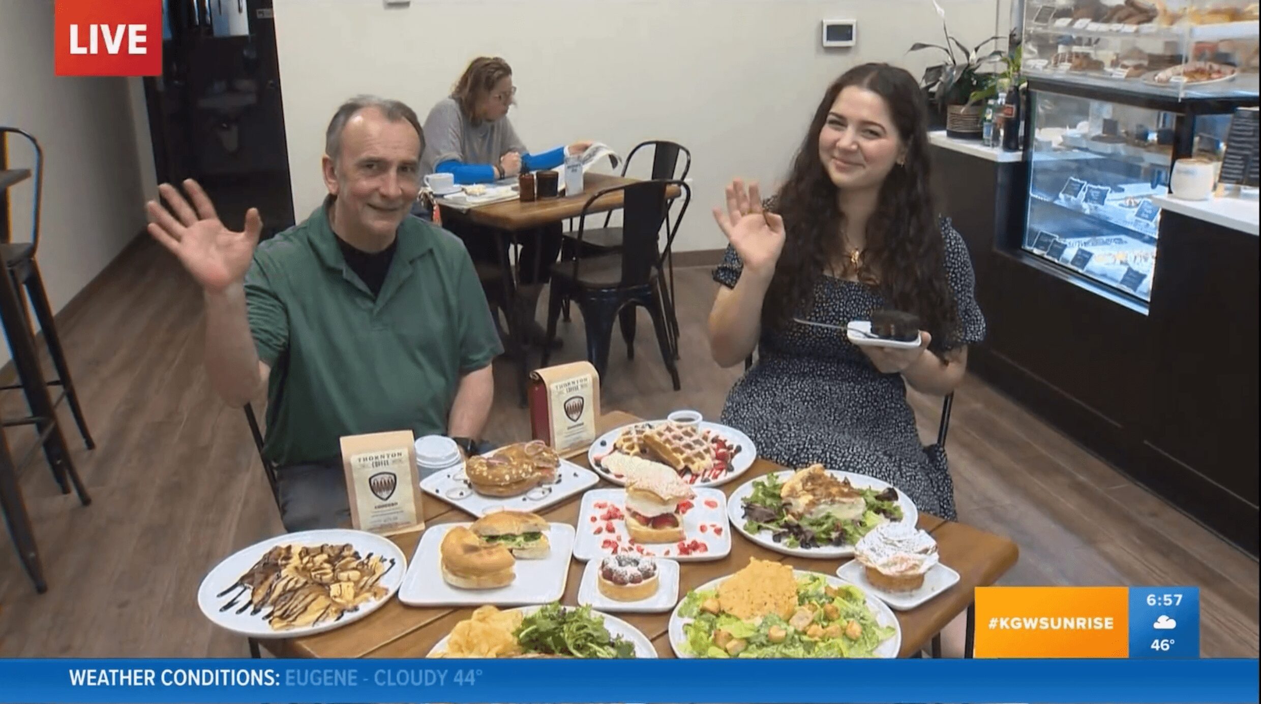 Two people sit at a table with various desserts and food, waving and smiling, in a cafe setting.