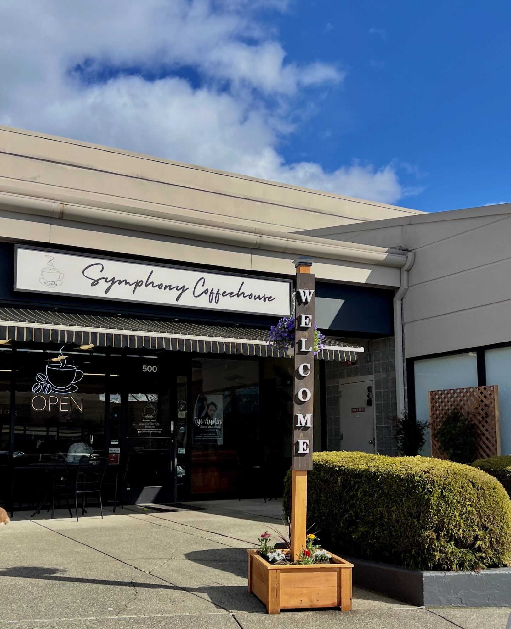 Coffee shop entrance with a sign reading 'Symphony Coffeehouse' and a vertical 'WELCOME' sign, bushes, and a clear blue sky.