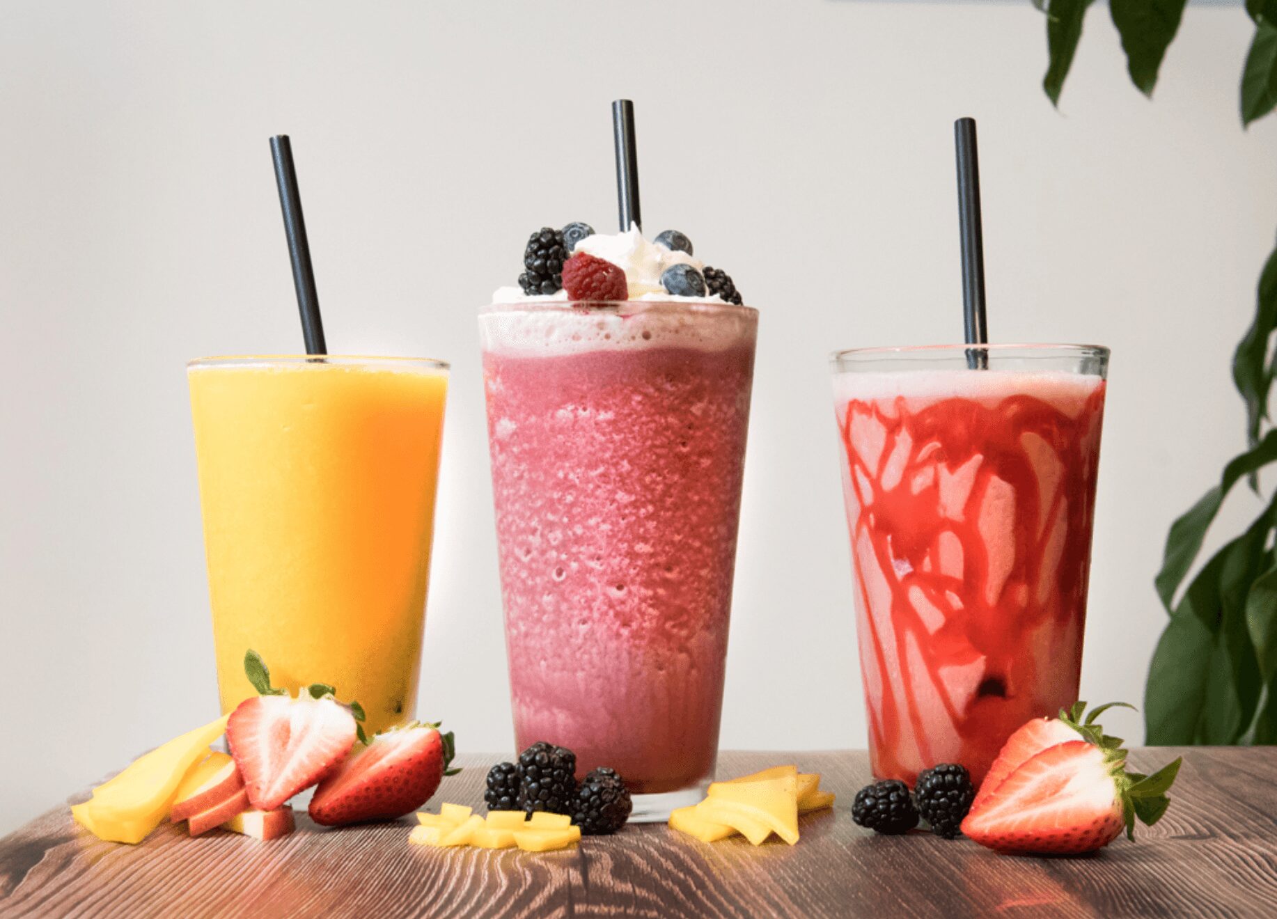 Three colorful smoothies on a wooden table with fruit garnishes and black straws, against a plain background.
