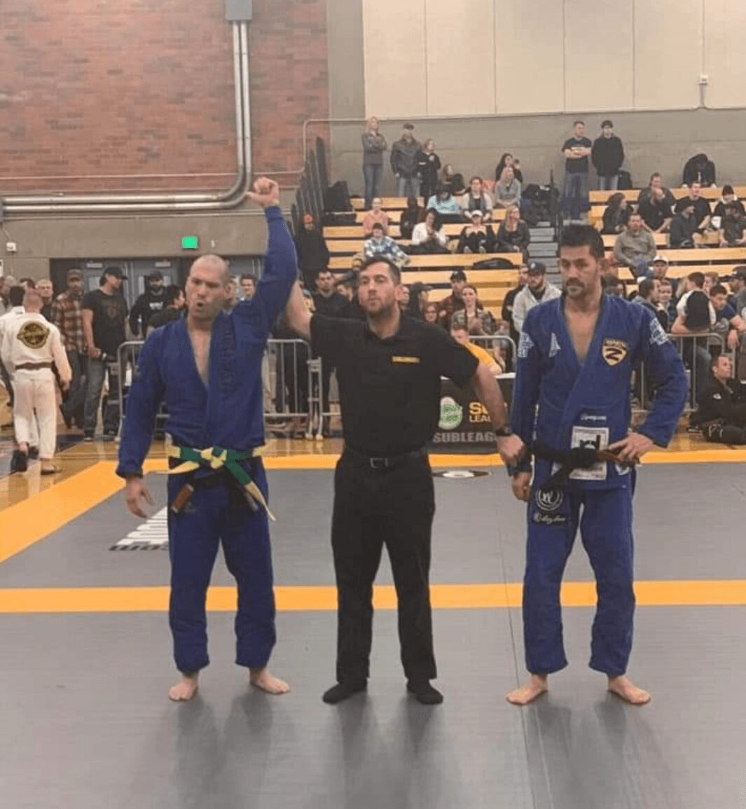 Two competitors in blue gis and a referee in black standing on a gray mat with yellow borders, in an indoor sports venue.
