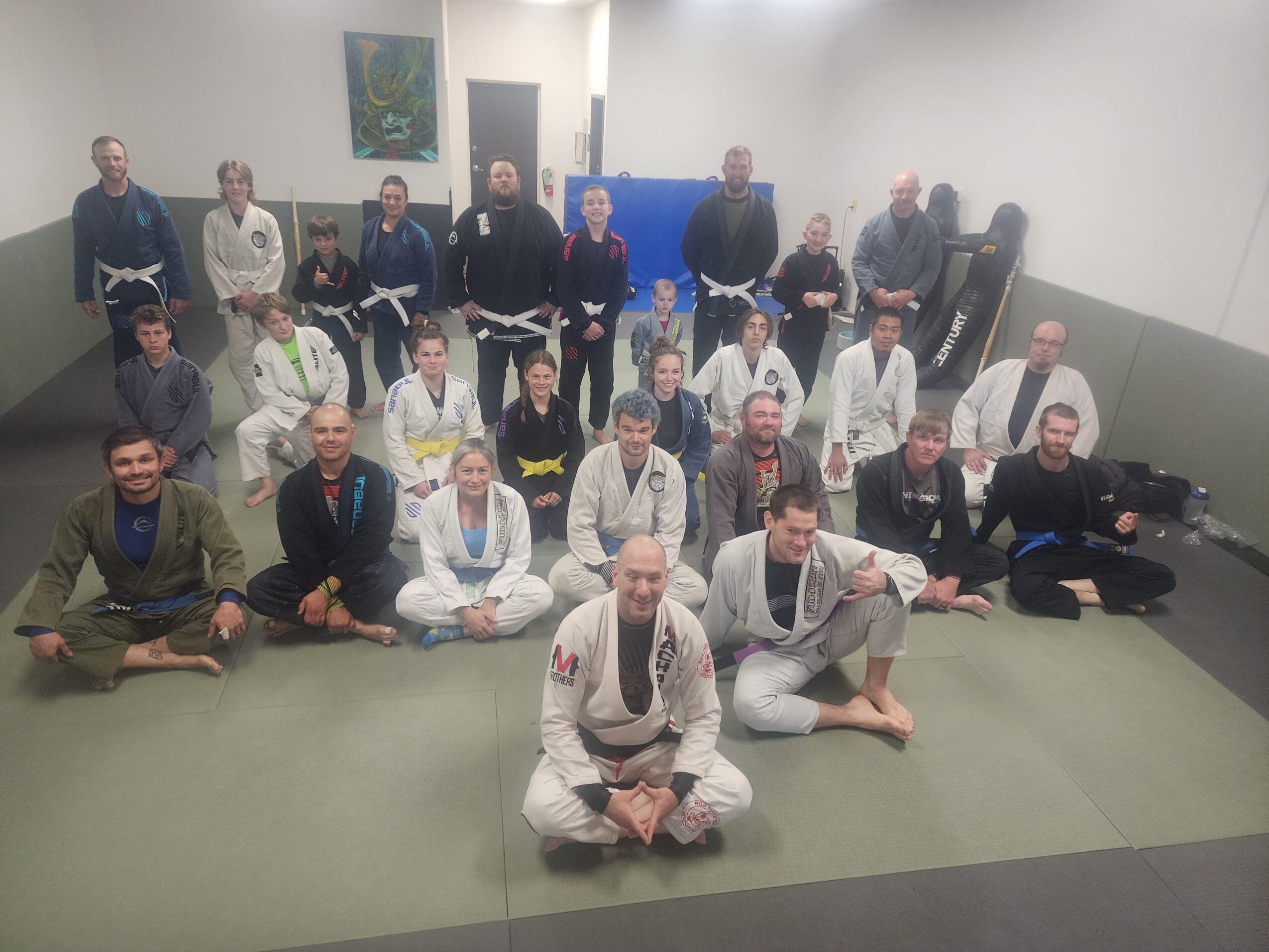 Group of people in martial arts uniforms and casual clothing in a dojo, some sitting and some standing, with mats on the floor.