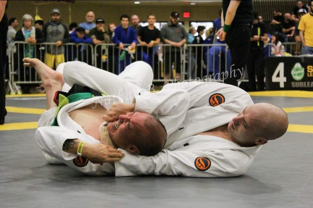 Two people in white martial arts uniforms grappling on the mat, with spectators watching in the background.