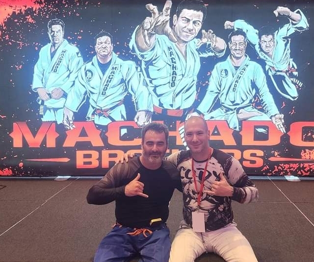 Two men sitting in front of a large poster of martial artists, one making a shaka sign, both smiling.