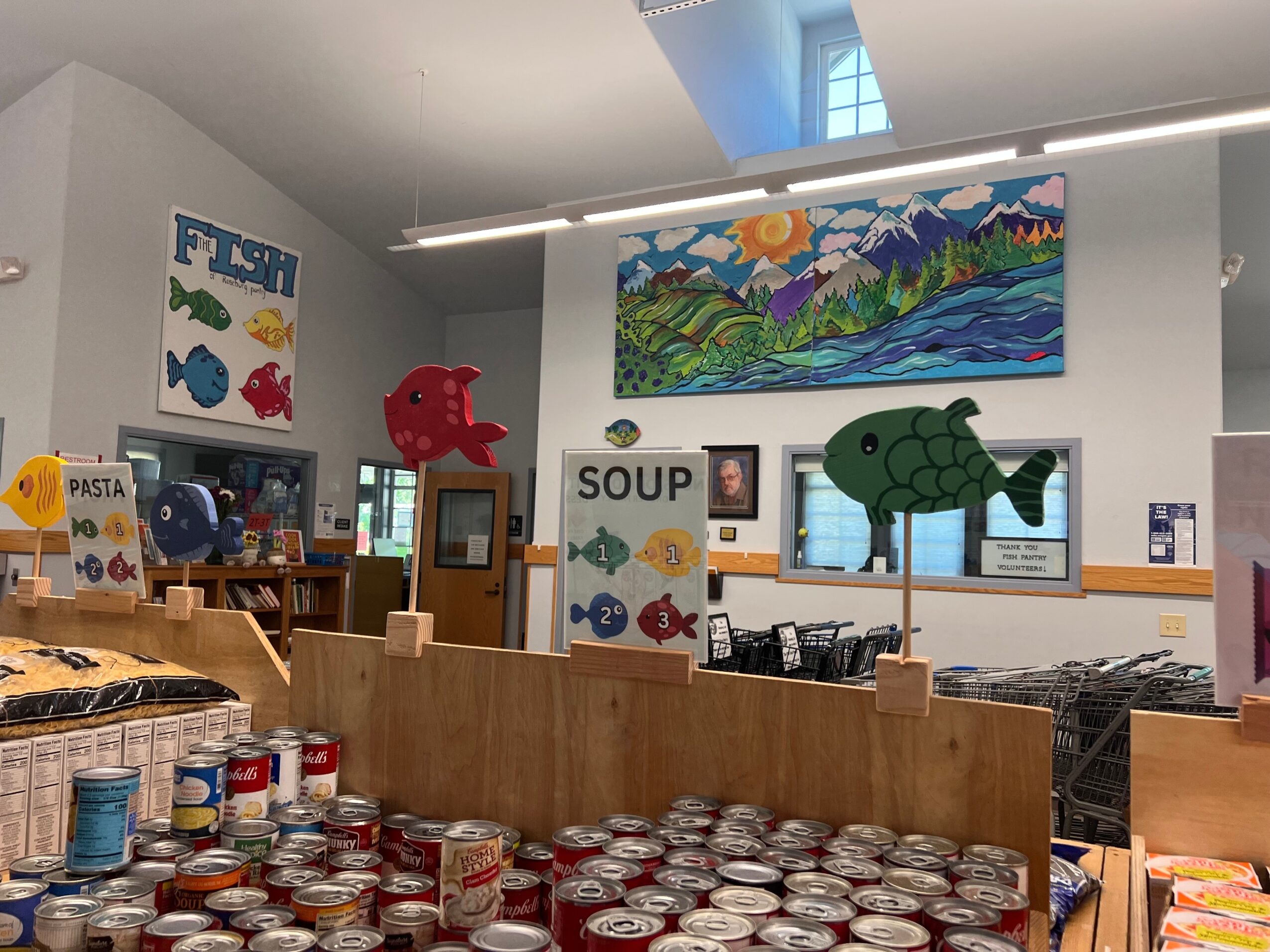 Indoor space with colorful fish decorations, a large landscape painting, and shelves with canned goods, possibly a library or community center.