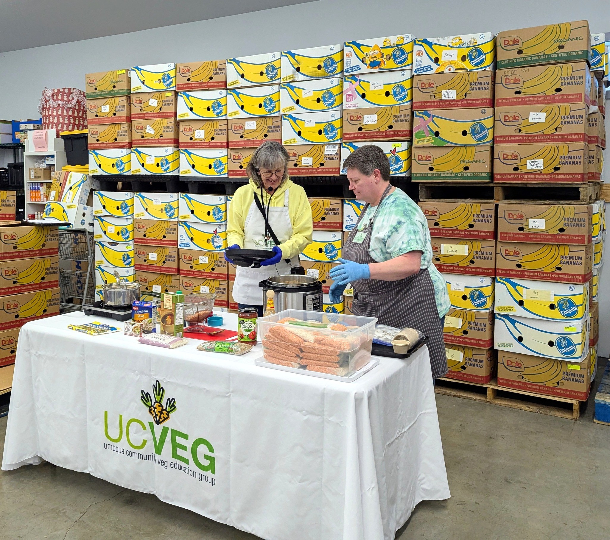 Two people work at a table with boxes of bananas in the background, wearing gloves and handling items. UCVG logo on tablecloth.