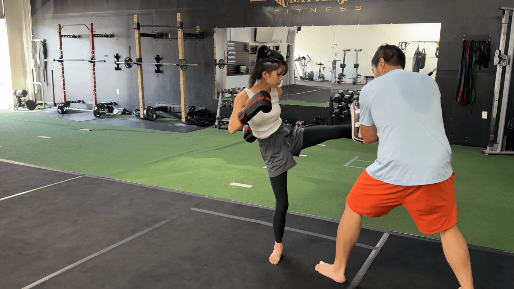A girl practicing martial arts with a man in a gym, girl performing a kick, man holding pads, gym equipment in background.