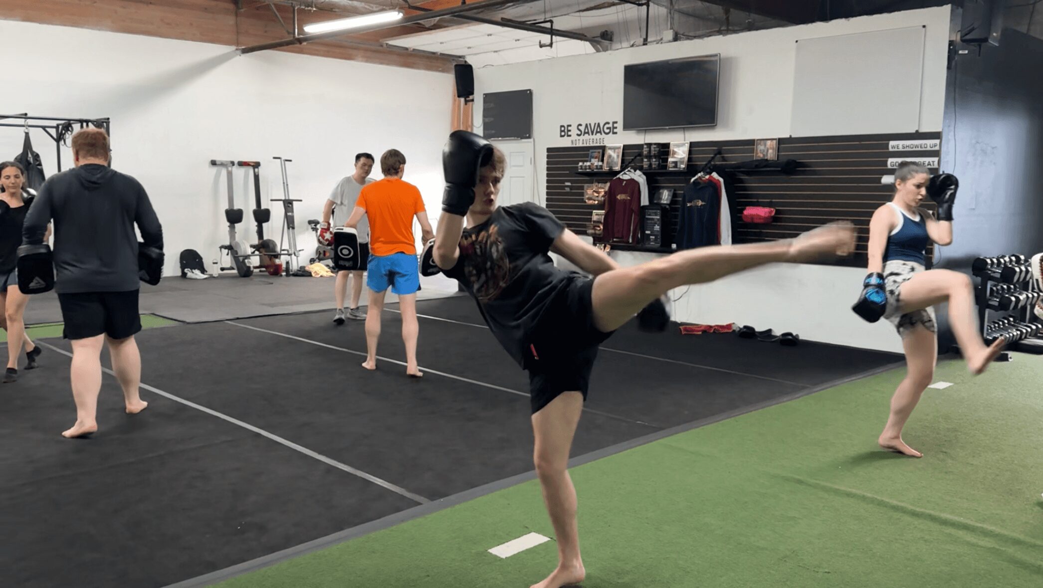 People training in a gym, some practicing kicks and stretches, with workout equipment in the background.