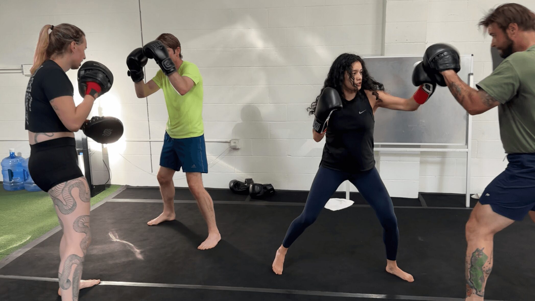 Four people practicing boxing in a gym, two wearing boxing gloves, in a training session.