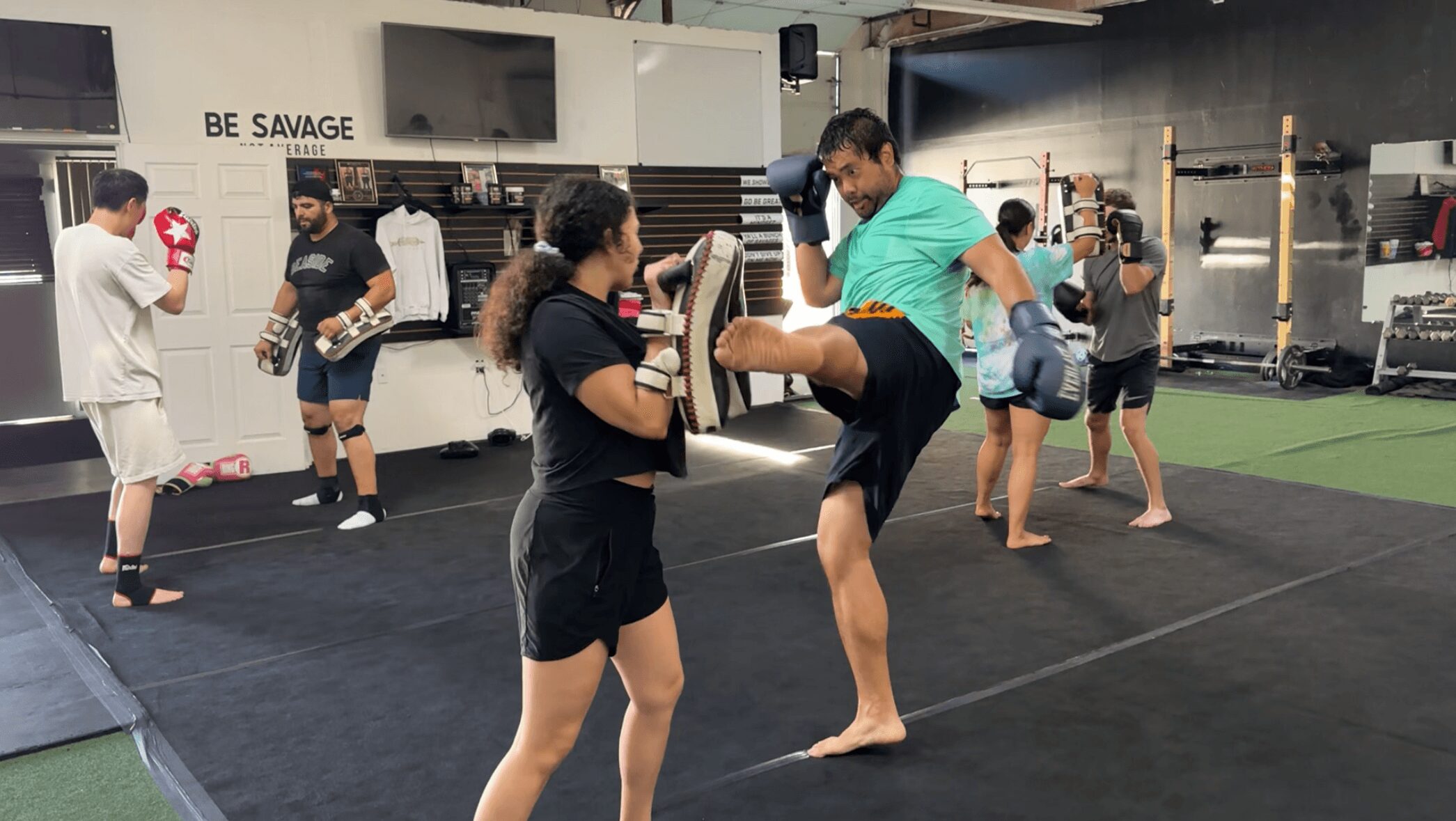 People practicing martial arts in a gym, some wearing gloves, others observing or training, with equipment in the background.
