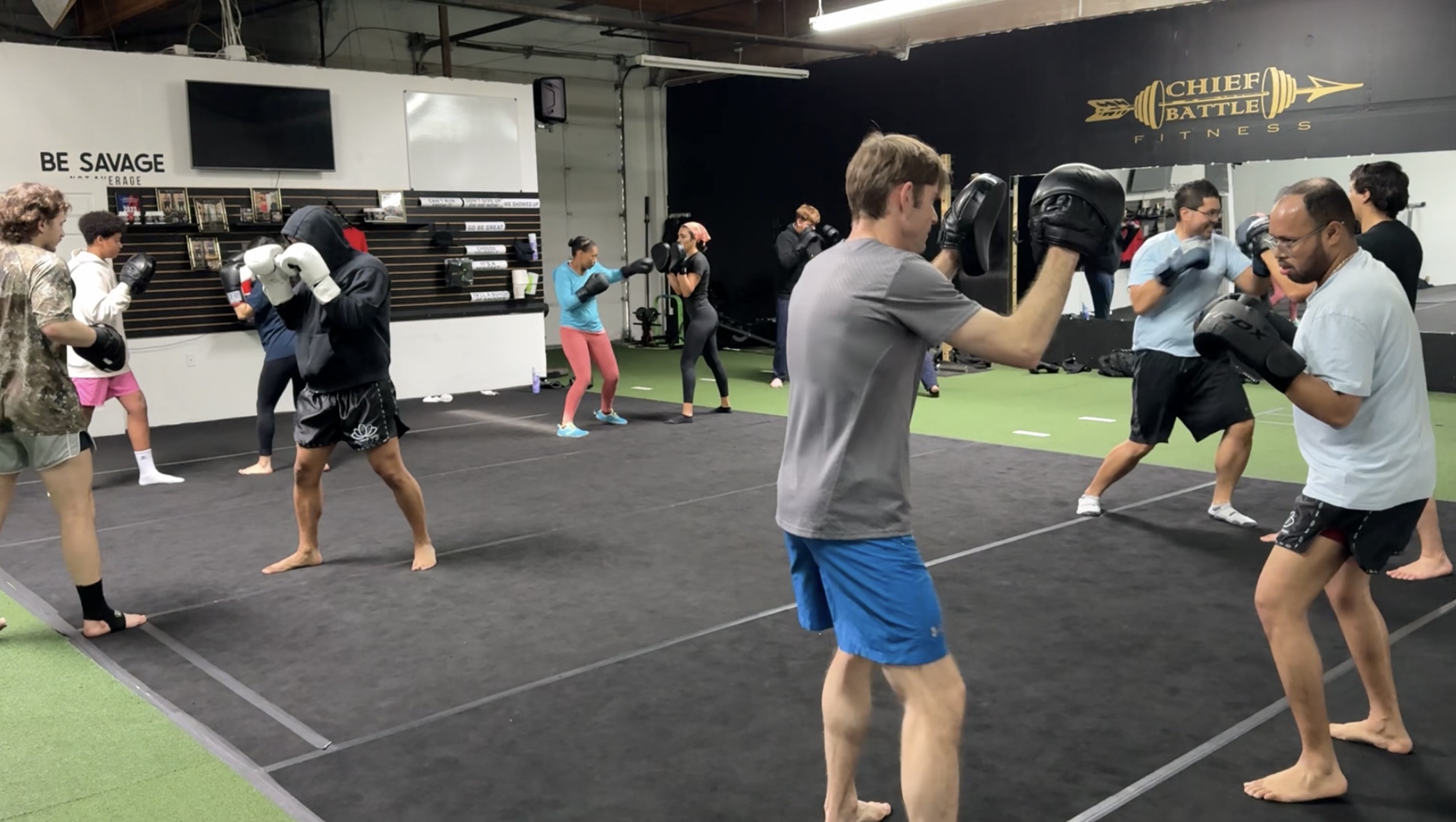 People practicing boxing in a gym with a black mat, some wearing gloves, others holding pads, and a sign reading 'BE SAVAGE'.