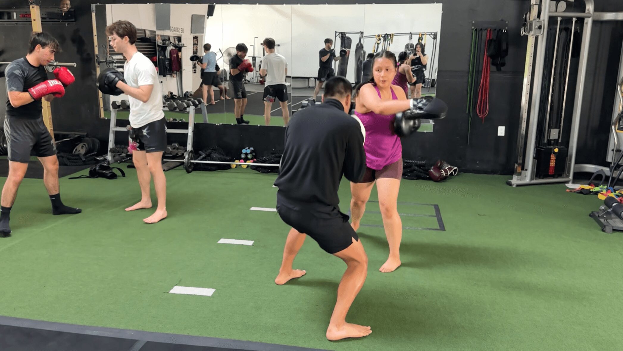 People practicing boxing in a gym with mirrors, mats, and equipment, some wearing gloves and shorts, others barefoot.
