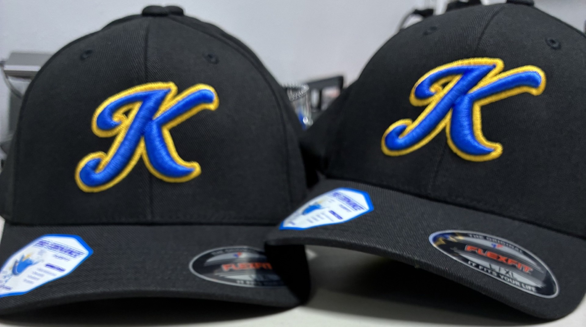 Two black caps with a blue and yellow embroidered letter 'K' on the front, placed side by side.