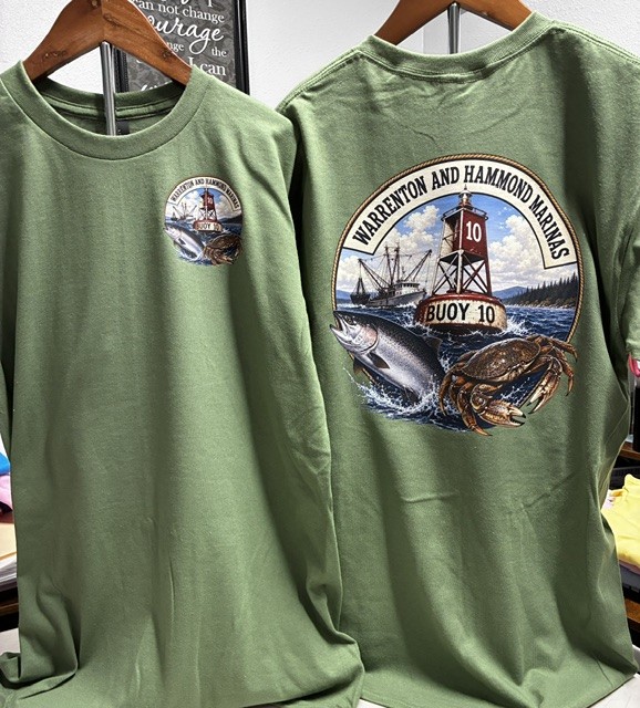 Two green long-sleeve shirts hanging on wooden hangers, one showing a small logo and the other a large circular graphic with a lighthouse, boat, and crab, and text about Warren and Hammond Marinas.