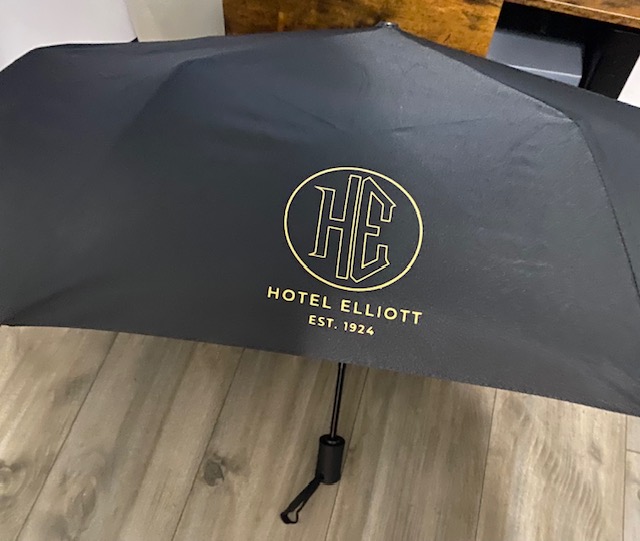 Black umbrella with gold logo and text for Hotel Elliott, standing on a wooden floor.
