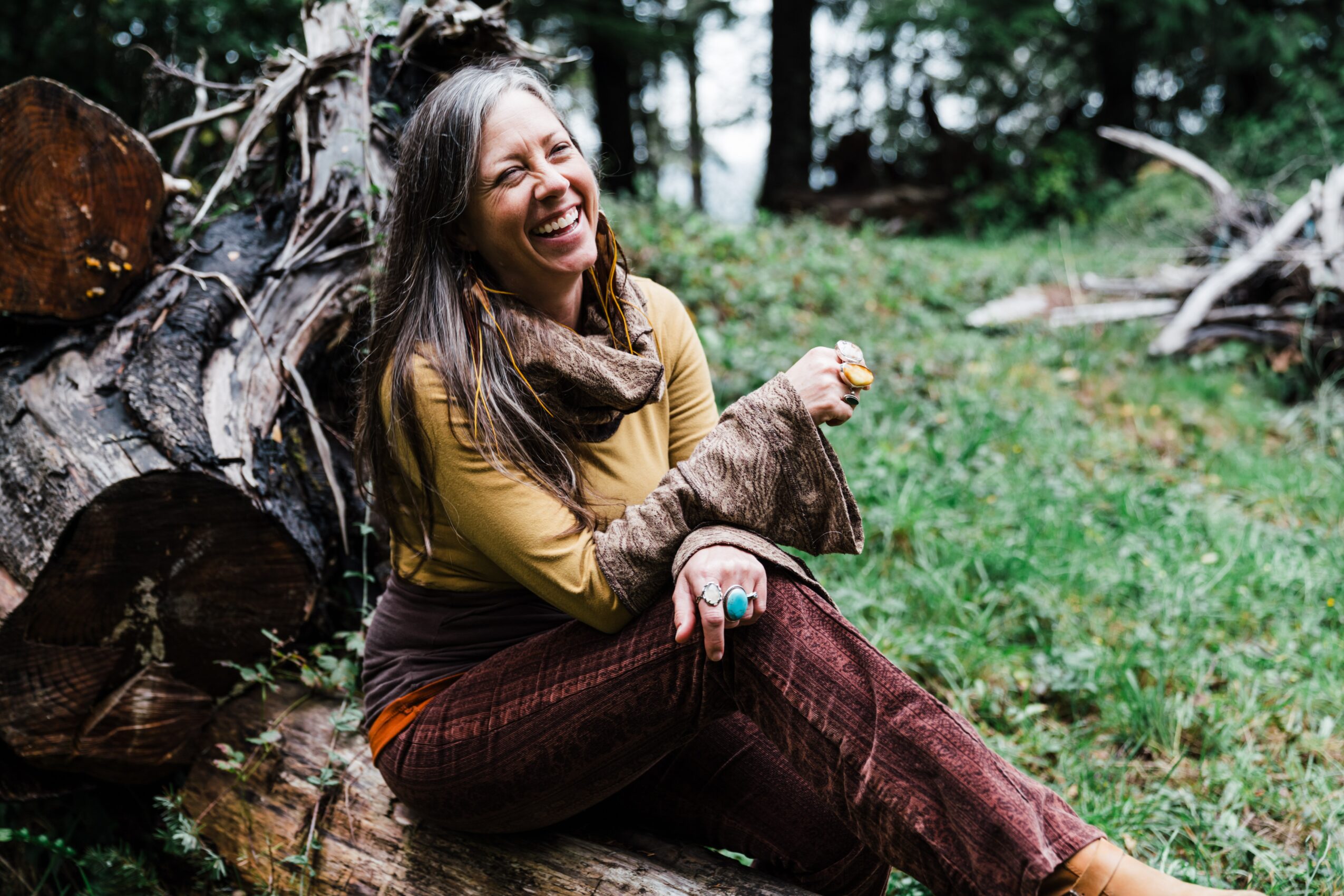 Woman sitting outdoors on grass, smiling and laughing, surrounded by trees and fallen logs.