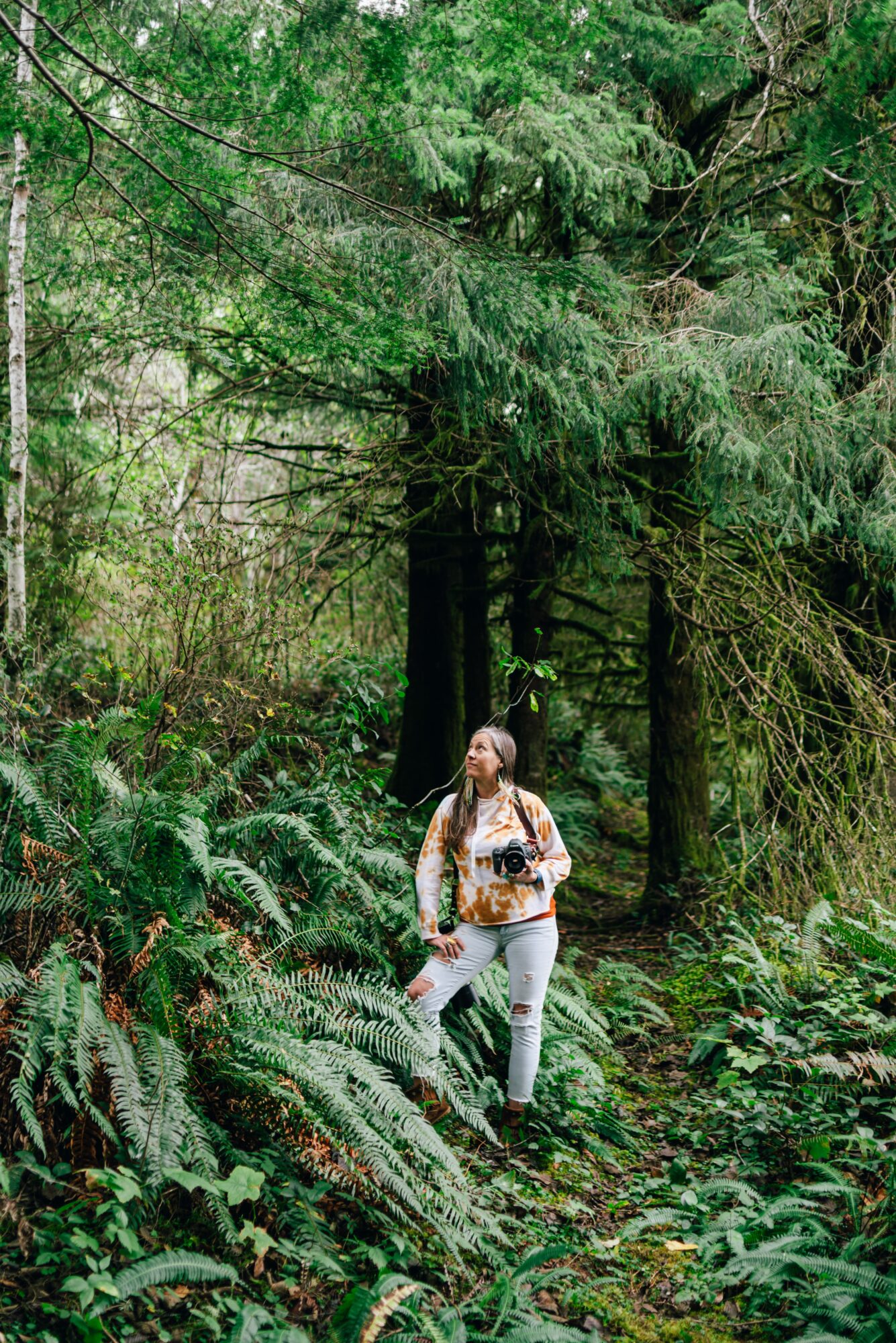 Woman walking in a dense forest with tall trees and lush green foliage, carrying a backpack and looking upward.