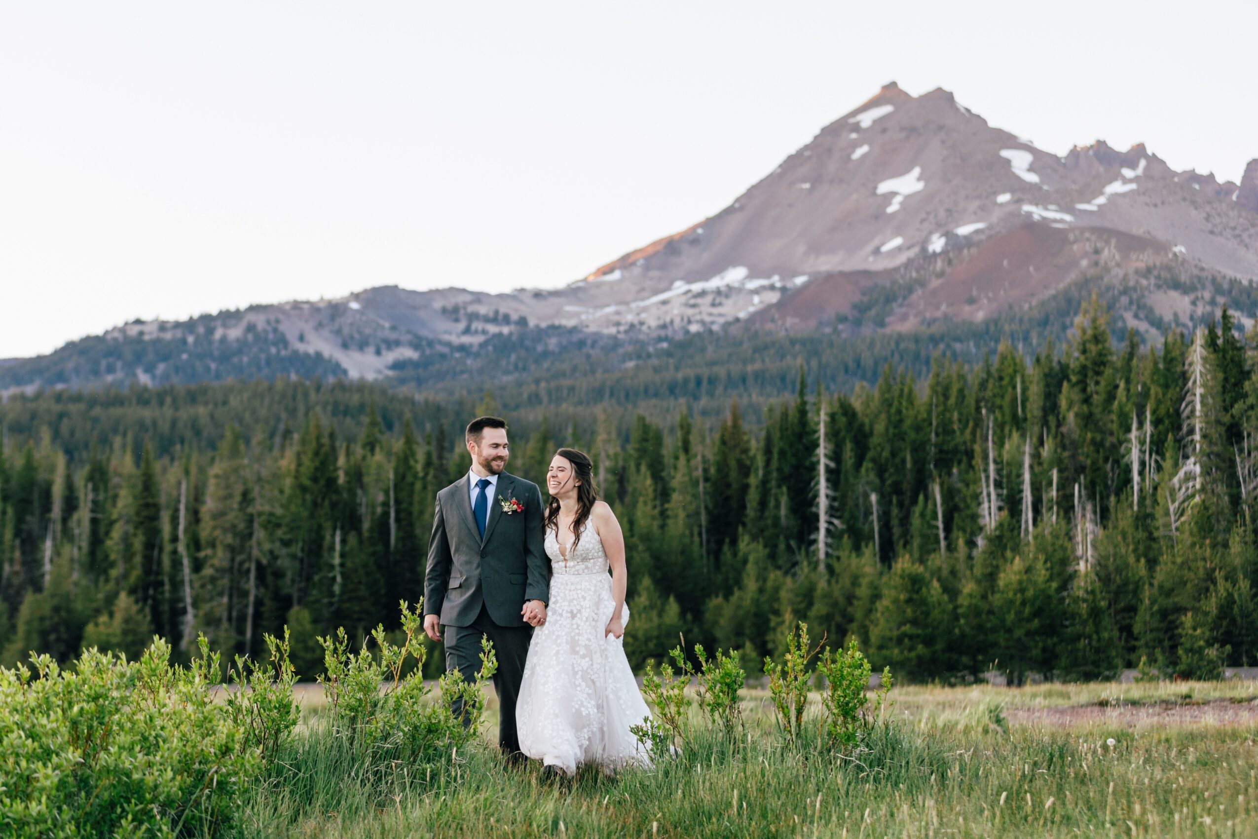 A couple in wedding attire walking hand in hand outdoors with a mountain and forest background.