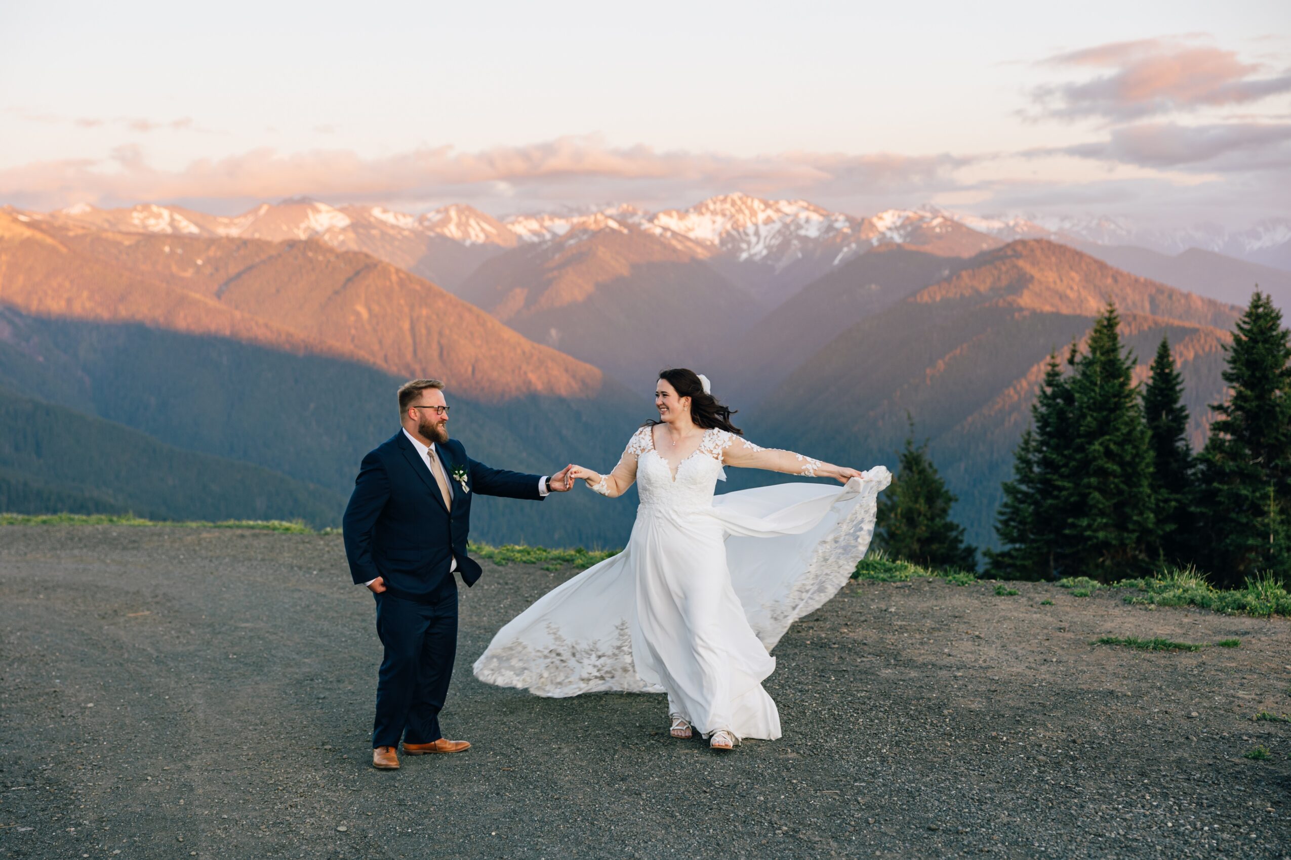 A couple in wedding attire holding hands outdoors with mountains and trees in the background, woman in a white dress, man in a suit.