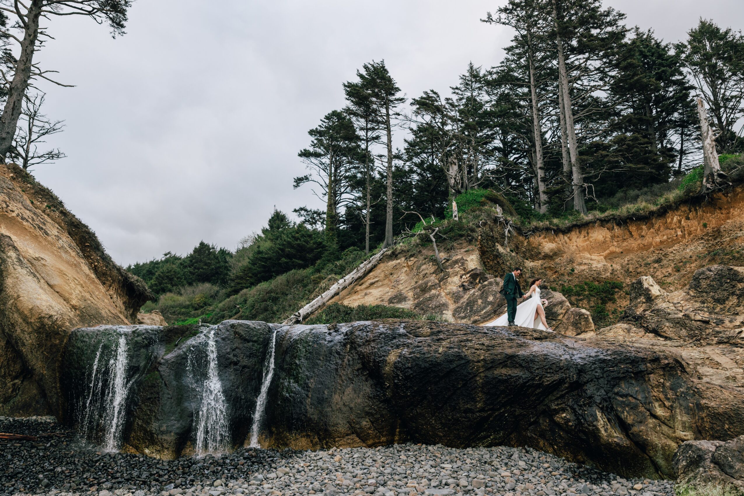 A person in a white dress stands on rocks near a small waterfall in a forested area under cloudy sky.
